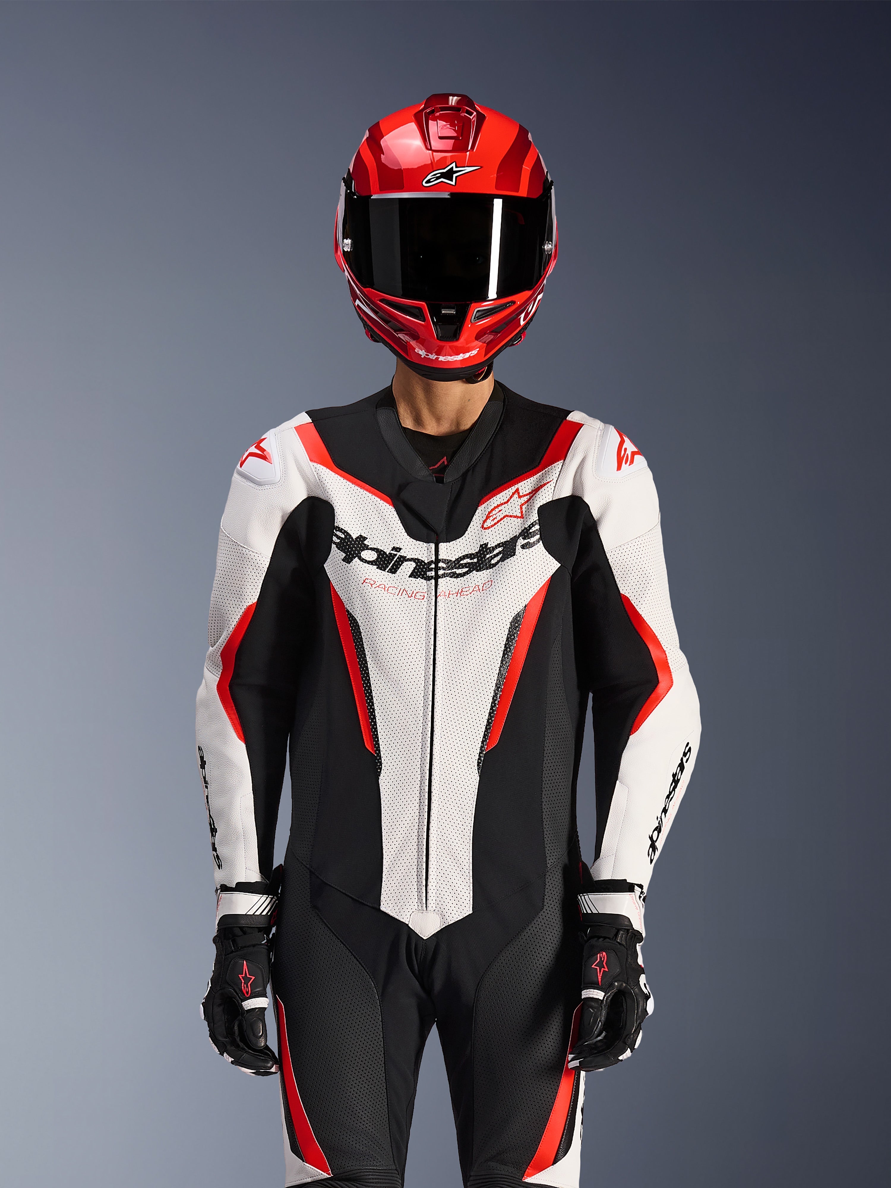 Alpinestars GP Force V2 Leren Pak - 1-delig, wit zwart rood fluo, gedragen met een rode integraalhelm en zwarte racehandschoenen, voorzien van geperforeerde leren panelen en opvallende Alpinestars-branding op de borst, professionele uitrusting voor motorraces