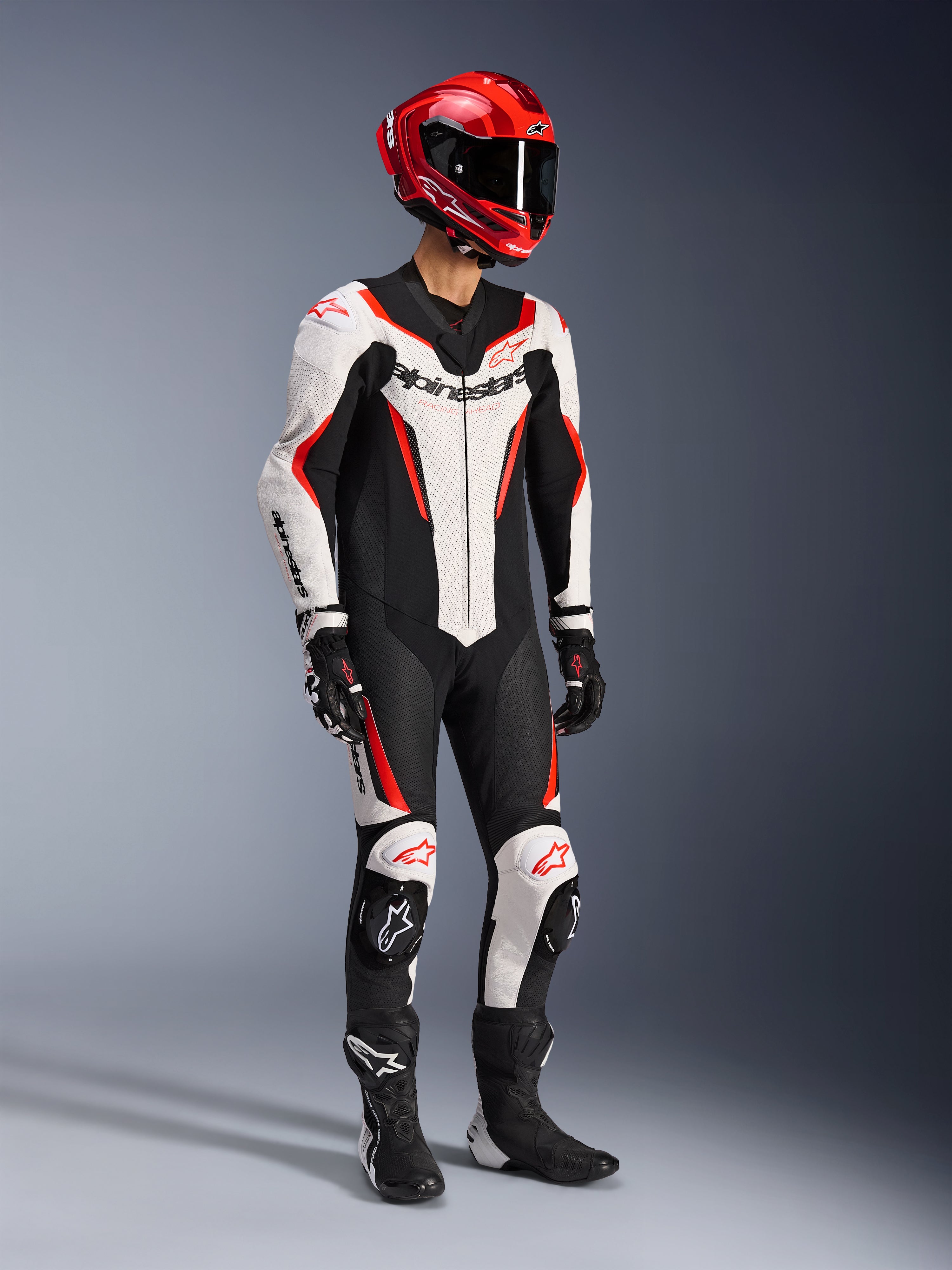 Alpinestars GP Force V2 Leren Pak 1-delig in Wit Zwart en Fluo Rood, studio-opname van een rijder in volledige race-uitrusting inclusief een rode helm, zwarte handschoenen en zwarte laarzen, met een aerodynamische runderleren constructie met opvallende branding en geïntegreerde kniesliders.
