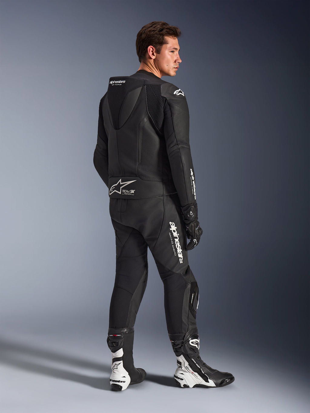 Mannelijk model draagt Alpinestars GP Force V2 2-delig leren motorpak in het zwart met witte logo's, driekwart achteraanzicht met de aerodynamische speedhump en Tech-Air Ready-branding, gecombineerd met zwart-witte racelaarzen en handschoenen, professionele sport- en circuitmotoruitrusting.