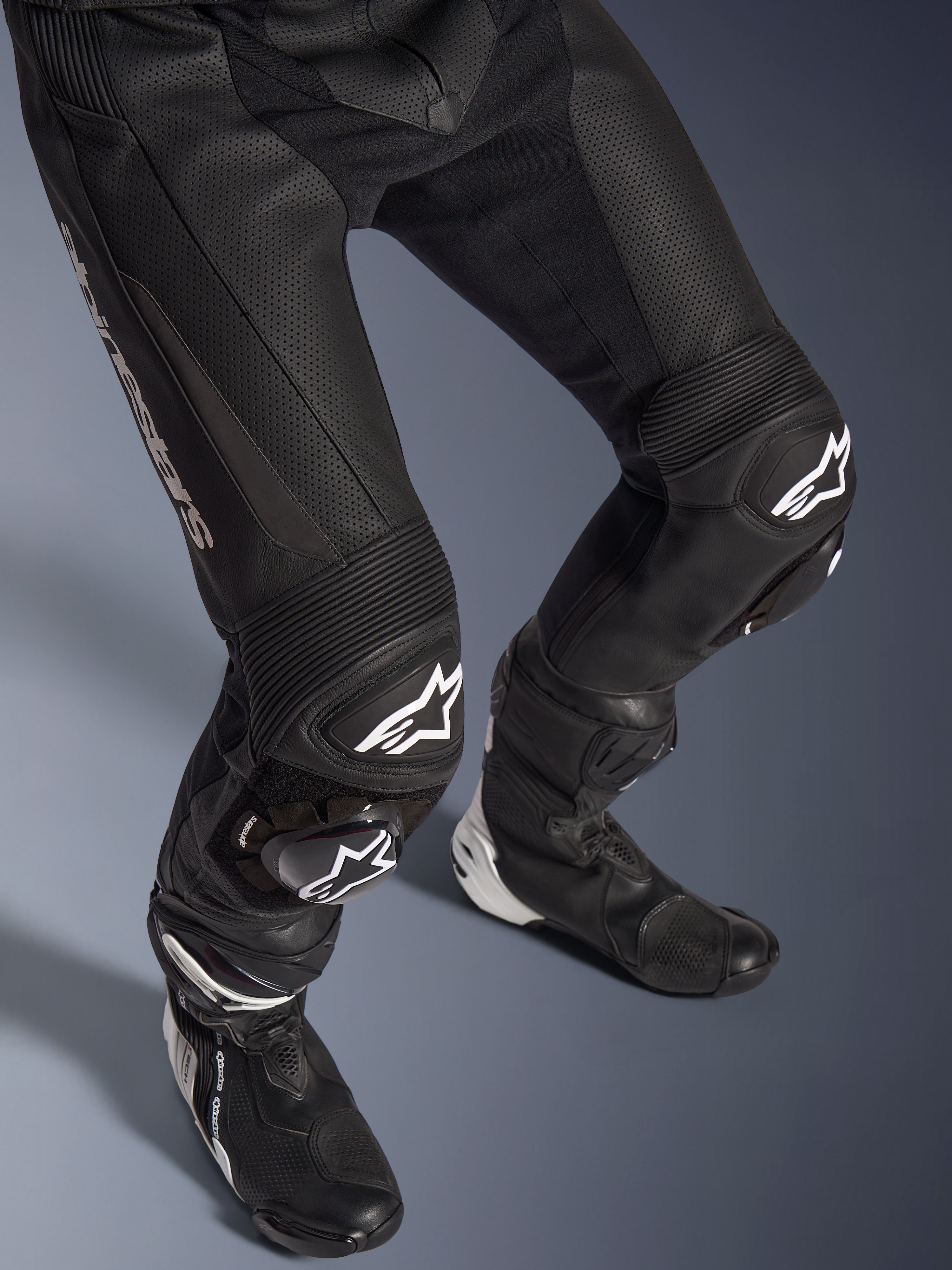 Alpinestars GP Force V2 2-delig leren pak, close-up van het onderlichaam inclusief broek en racelaarzen, zwart met witte logo's, geperforeerde leren constructie, accordeon stretchpanelen op de knieën en beschermende sliders, professionele sportieve motoruitrusting