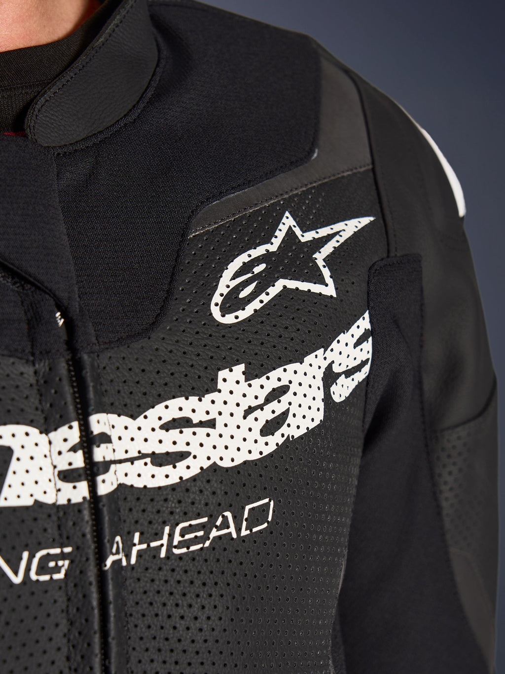 Alpinestars GP Force V2 2-delig leren pak, racing/sport motorpak, zwart, close-up van de geperforeerde lederen borst met wit Alpinestars-logo en branding, nadruk op ventilatieopeningen en getextureerde constructie.