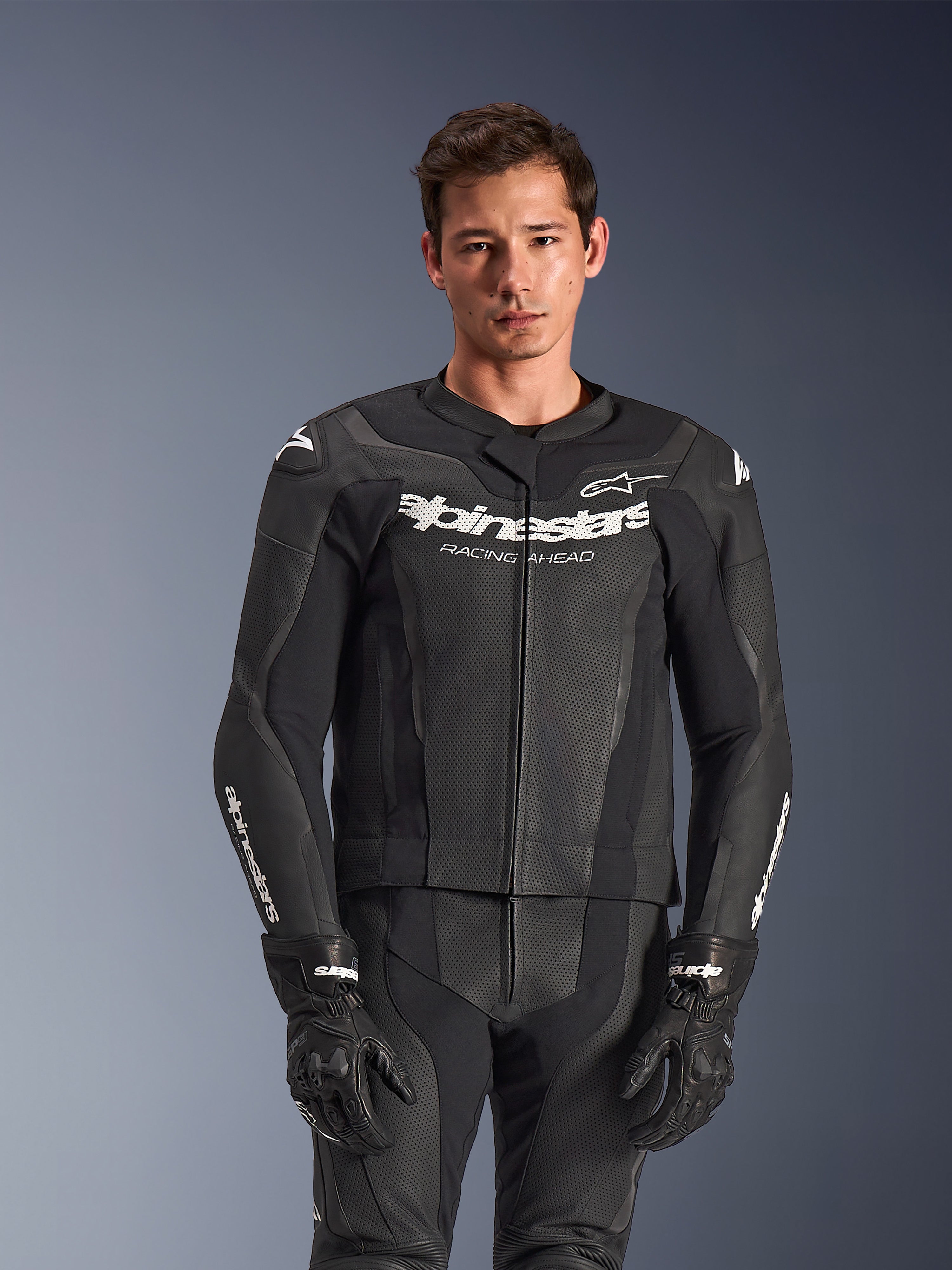 Alpinestars GP Force V2 2-delig leren pak, zwart, gedragen door model, met geperforeerde leren panelen en witte 'Alpinestars Racing Ahead' branding op de borst, gecombineerd met zwarte handschoenen, professionele sportieve motoruitrusting