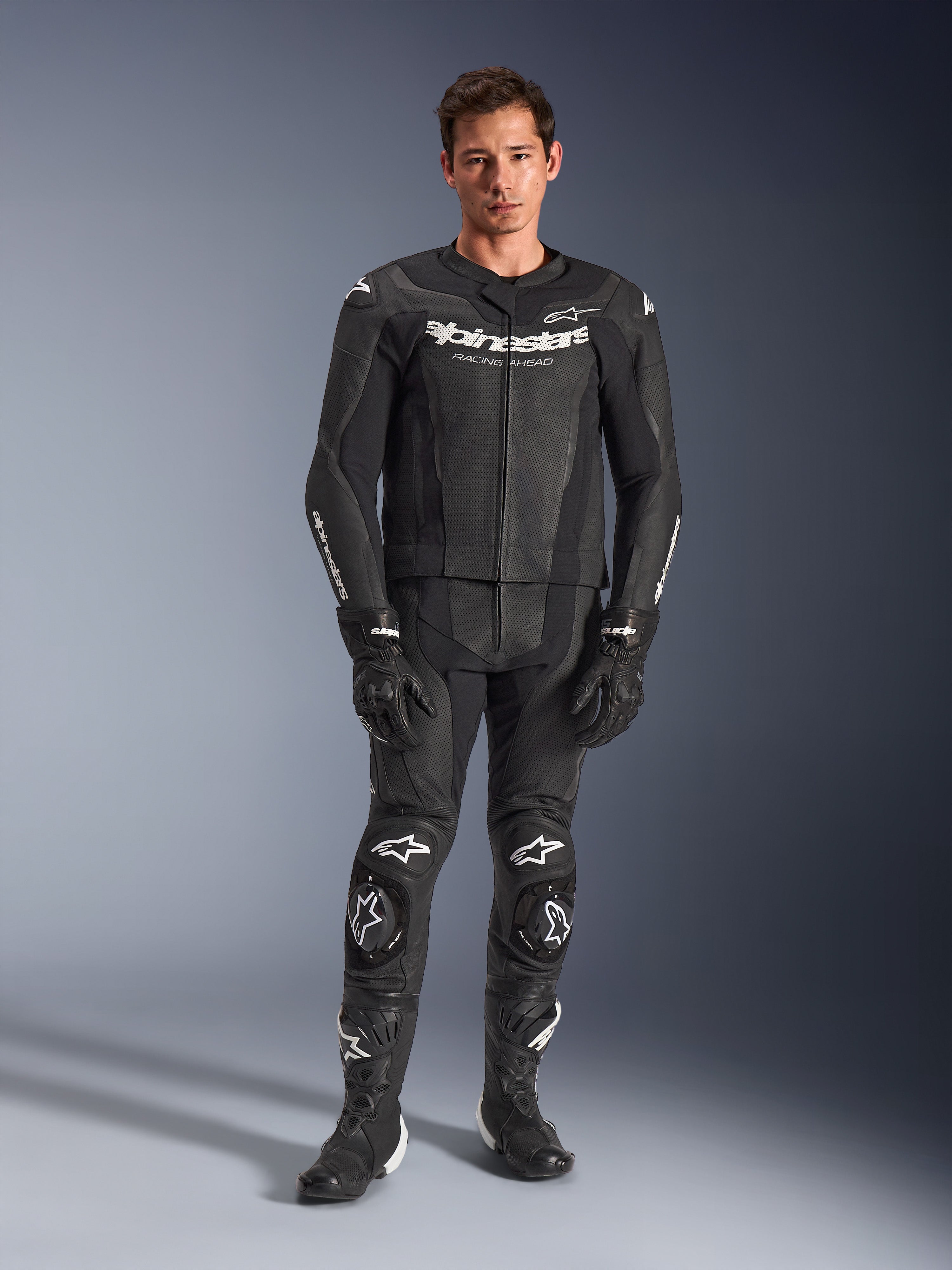 Alpinestars GP Force V2 tweedelig leren pak, zwart met witte logo's, volledig motorracepak, hoogwaardige leren constructie met verstevigde kniesliders en geperforeerde panelen, gedragen met zwarte handschoenen en laarzen, studio-opname in volledige lengte