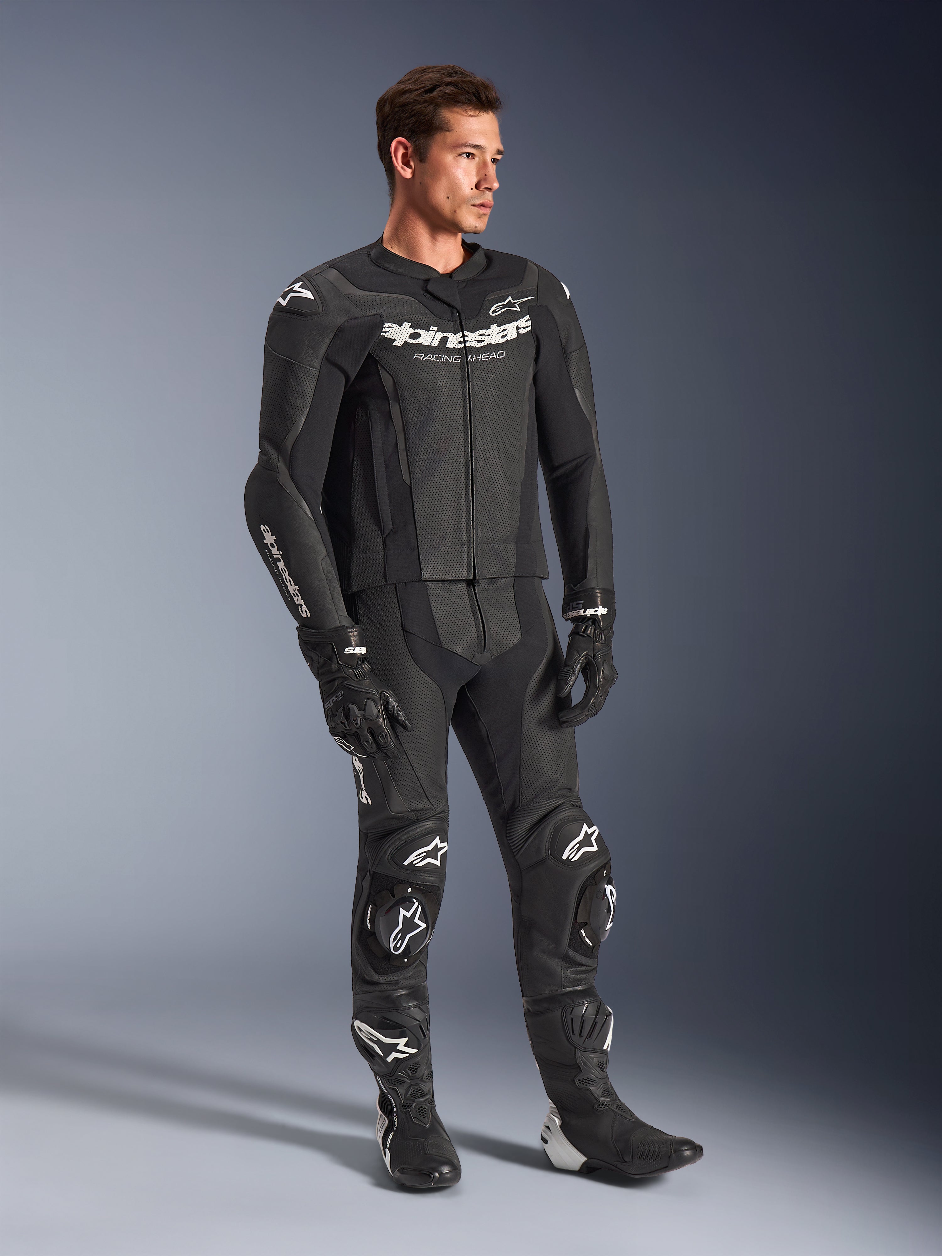 Alpinestars GP Force V2 2-delig leren pak, zwart, gedragen door een mannelijk model met bijpassende zwarte racehandschoenen en laarzen, voorzien van witte Alpinestars-branding, geperforeerde leren panelen voor ventilatie en geïntegreerde kniesliders, professionele motorrace-uitrusting