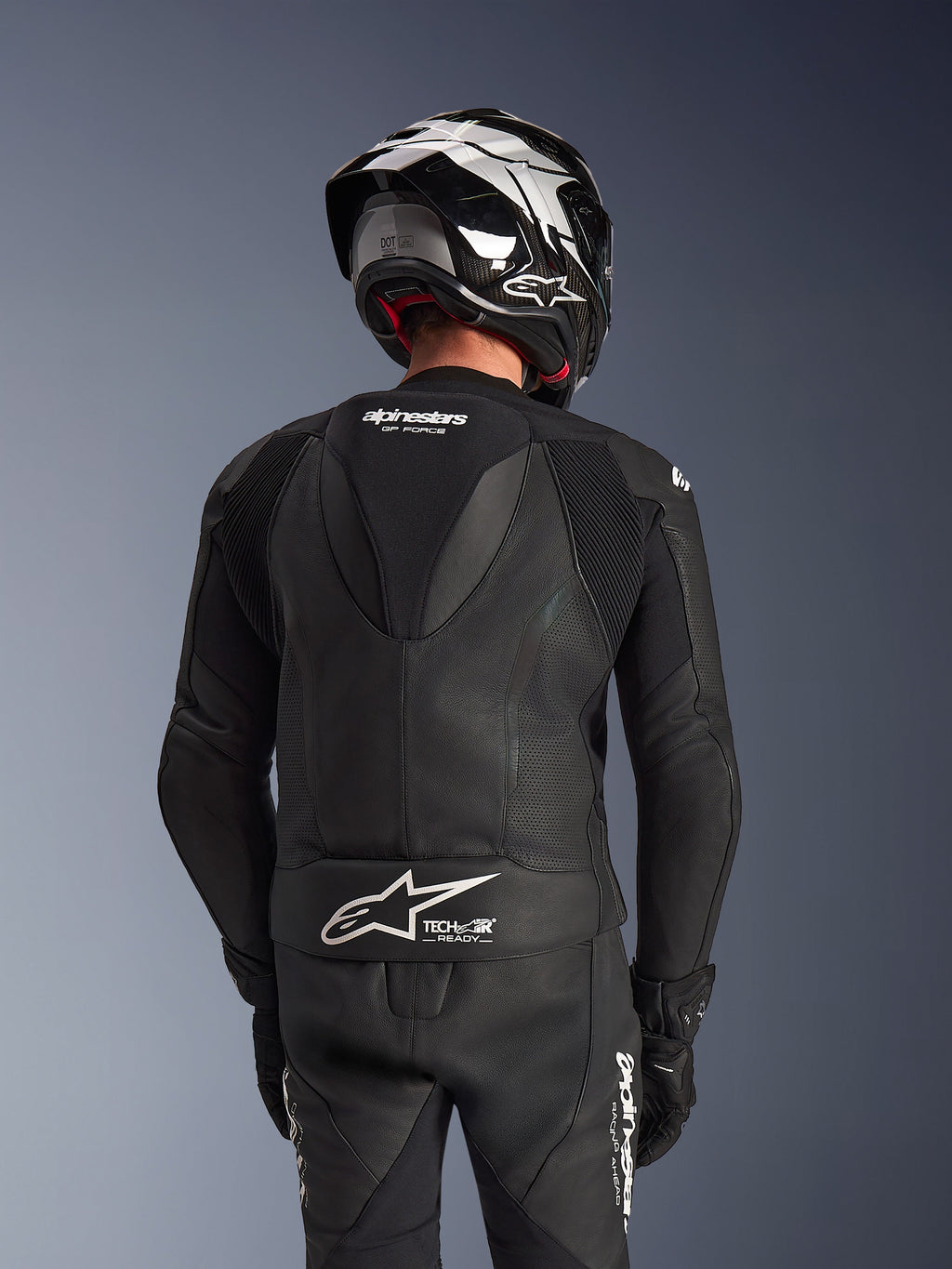 Alpinestars GP Force V2 2-delig leren pak, zwart, achteraanzicht van de rijder met aerodynamische speedhump, geperforeerde leren panelen en accordeon-stretchzones op de schouders, Tech-Air Ready-branding op de onderrug, gedragen met bijpassende racehelm en handschoenen.