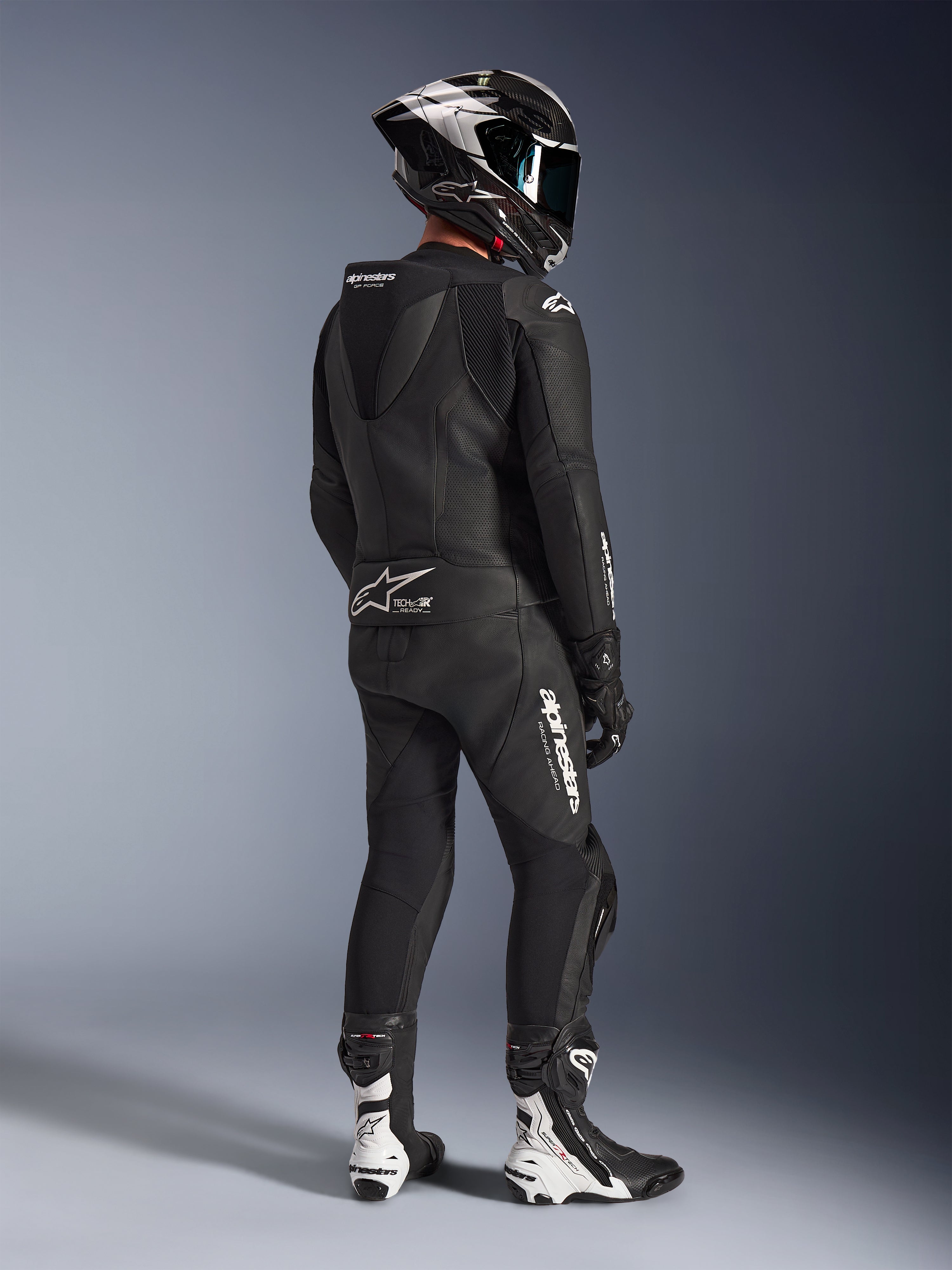 Alpinestars GP Force V2 2-delig leren pak, zwart, achteraanzicht van motorrijder in volledige race-uitrusting, met aerodynamische speedhump, witte logo's en geperforeerd leer, gecombineerd met een carbon helm en zwart-witte racelaarzen
