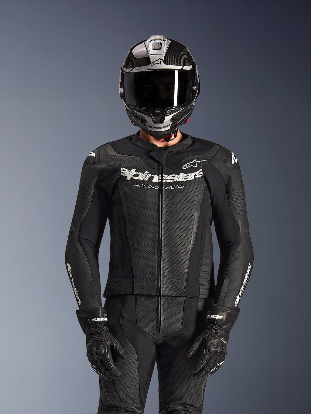 Rijder in Alpinestars GP Force V2 2-delig leren pak in het zwart, geperforeerde leren constructie met wit 'Racing Ahead' logo op de borst, zilveren en carbonfiber integraalhelm, zwarte handschoenen, professionele sport- en circuituitrusting