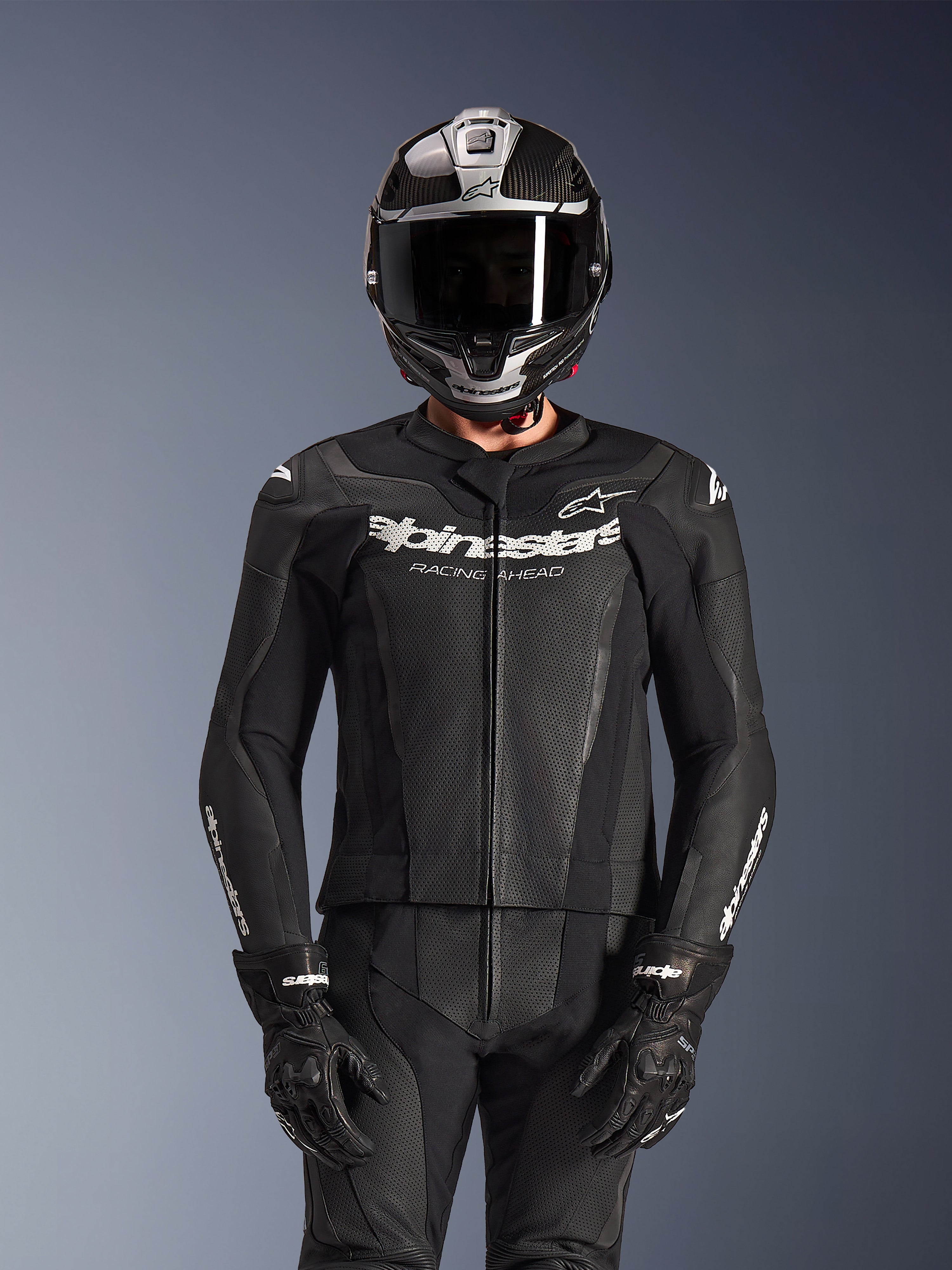 Rijder in Alpinestars GP Force V2 2-delig leren pak in het zwart, geperforeerde leren constructie met wit 'Racing Ahead' logo op de borst, zilveren en carbonfiber integraalhelm, zwarte handschoenen, professionele sport- en circuituitrusting