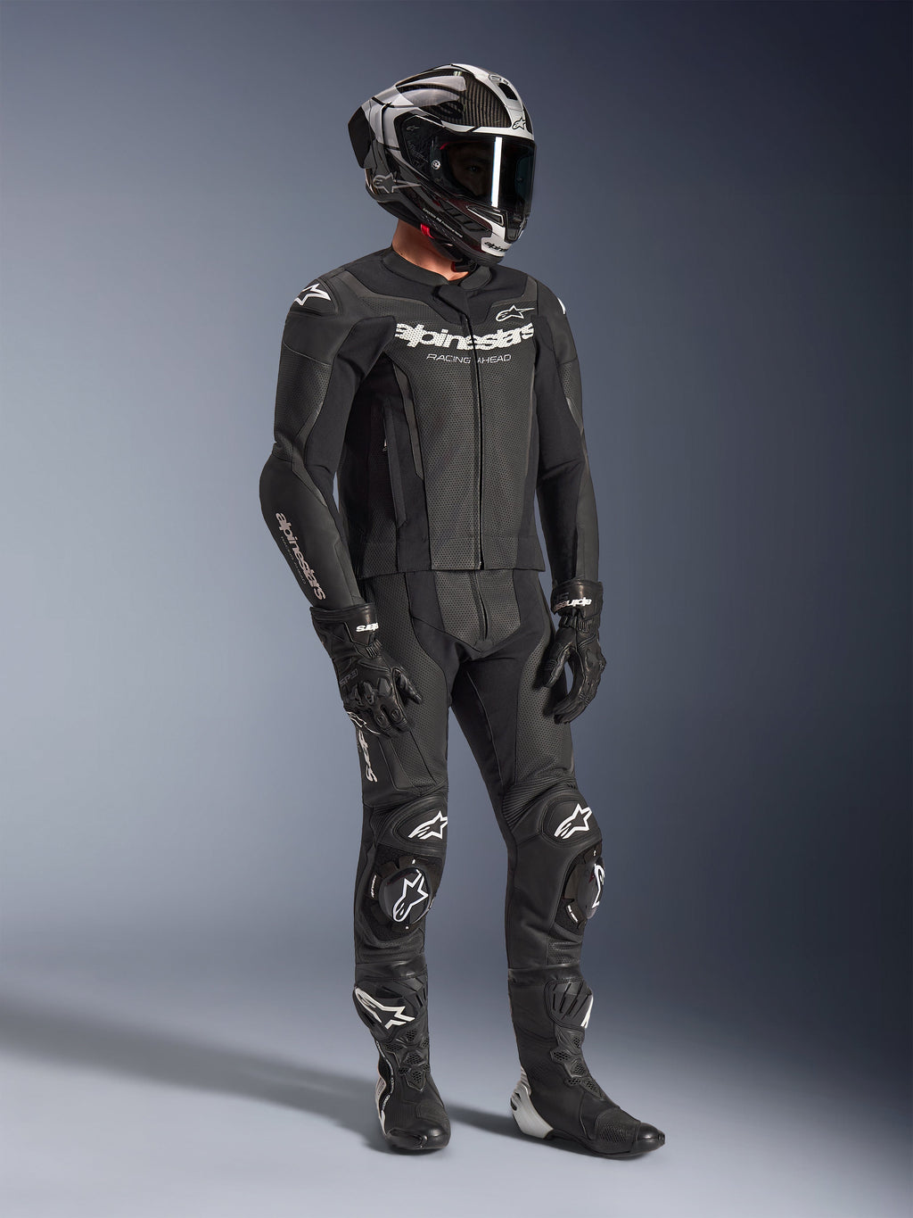 Alpinestars GP Force V2 2-delig leren pak, zwart motorracepak, gedragen met integraalhelm, handschoenen en laarzen, geperforeerde leren constructie, aerodynamische speedhump en witte Alpinestars-logo's op de borst, armen en knieën
