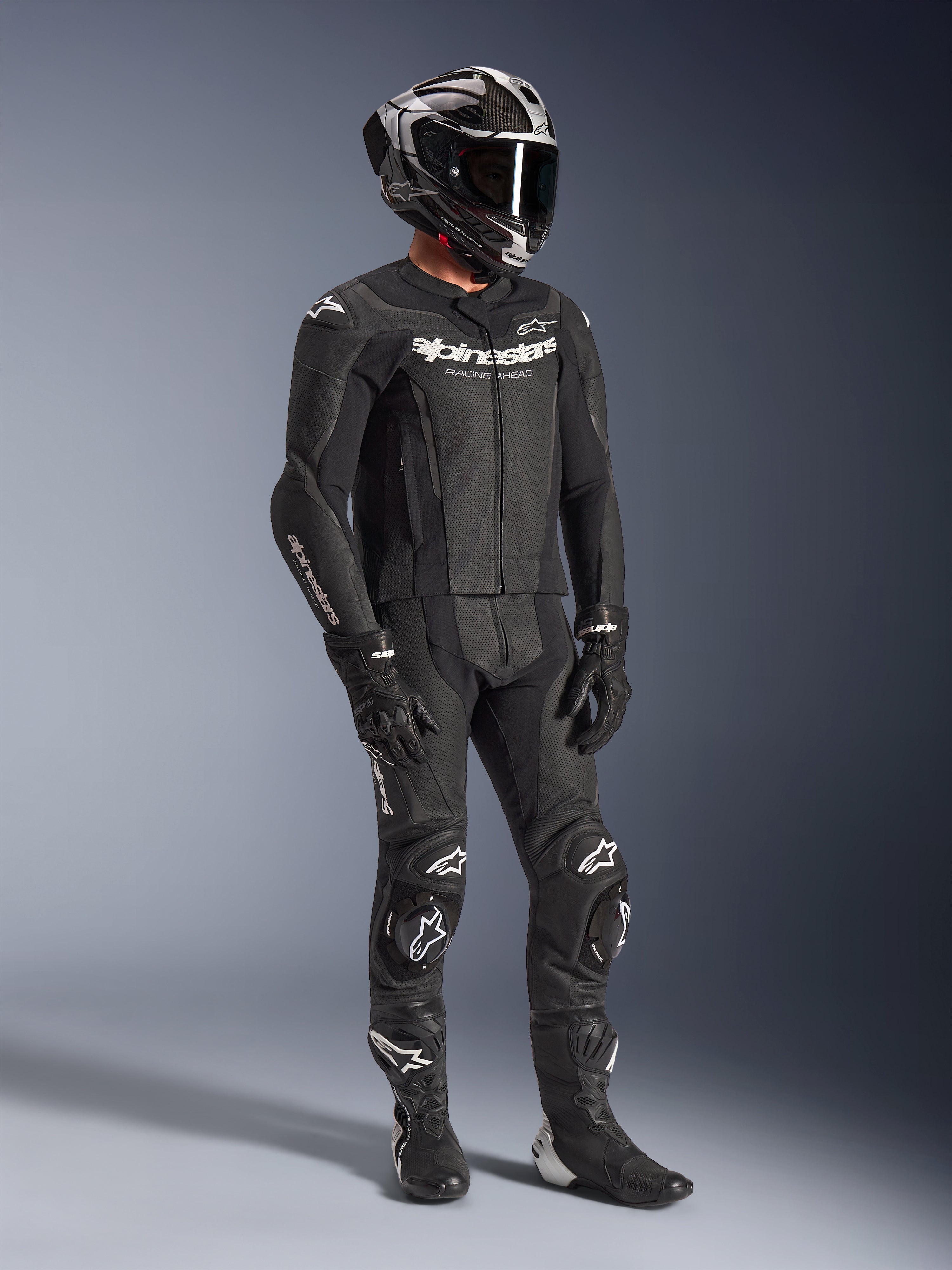 Alpinestars GP Force V2 2-delig leren pak, zwart motorracepak, gedragen met integraalhelm, handschoenen en laarzen, geperforeerde leren constructie, aerodynamische speedhump en witte Alpinestars-logo's op de borst, armen en knieën