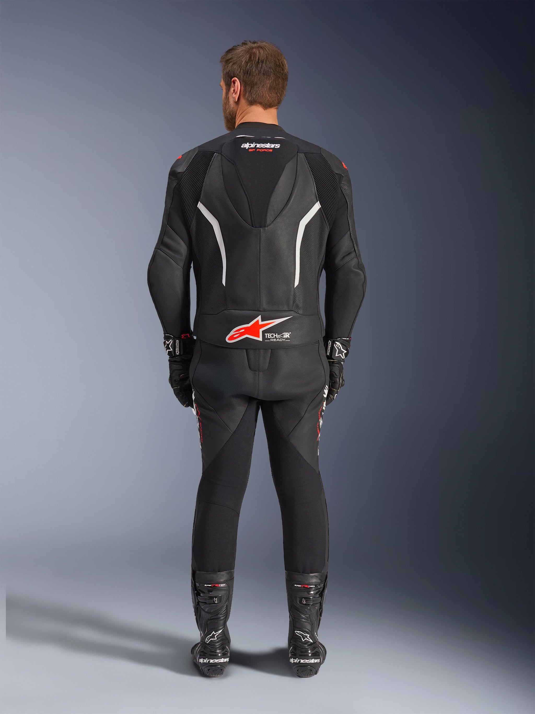 Achteraanzicht van een rijder in het Alpinestars GP Force V2 tweedelig leren pak, zwart met witte en rode accenten, opvallende Alpinestars en Tech-Air Ready branding op de rug, gestyled met bijpassende zwarte racehandschoenen en laarzen