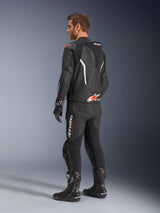 Alpinestars GP Force V2 tweedelig leren pak, zwart met witte en Red Fluo accenten, driekwart achteraanzicht op model, met een aerodynamische speed hump, geperforeerde leren panelen en opvallende Alpinestars branding, gedragen met zwarte racelaarzen en handschoenen.