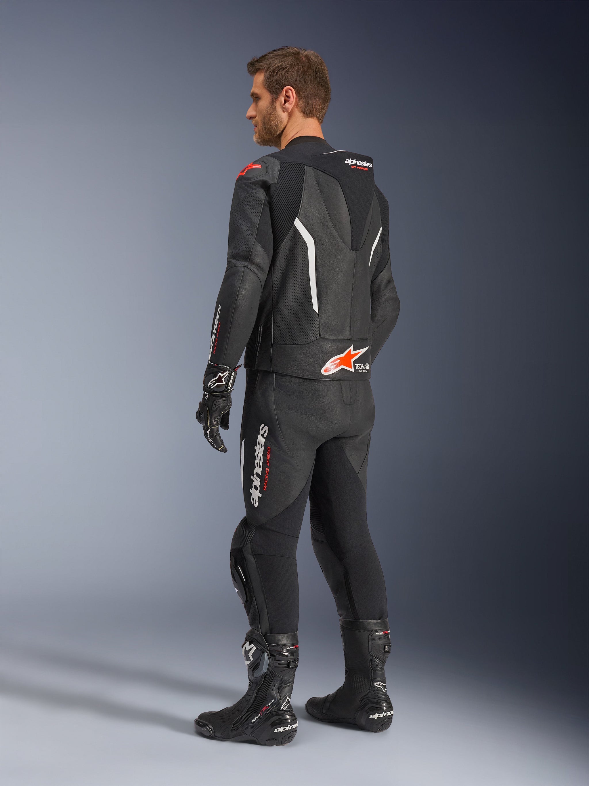 Alpinestars GP Force V2 tweedelig leren pak, zwart met witte en Red Fluo accenten, driekwart achteraanzicht op model, met een aerodynamische speed hump, geperforeerde leren panelen en opvallende Alpinestars branding, gedragen met zwarte racelaarzen en handschoenen.
