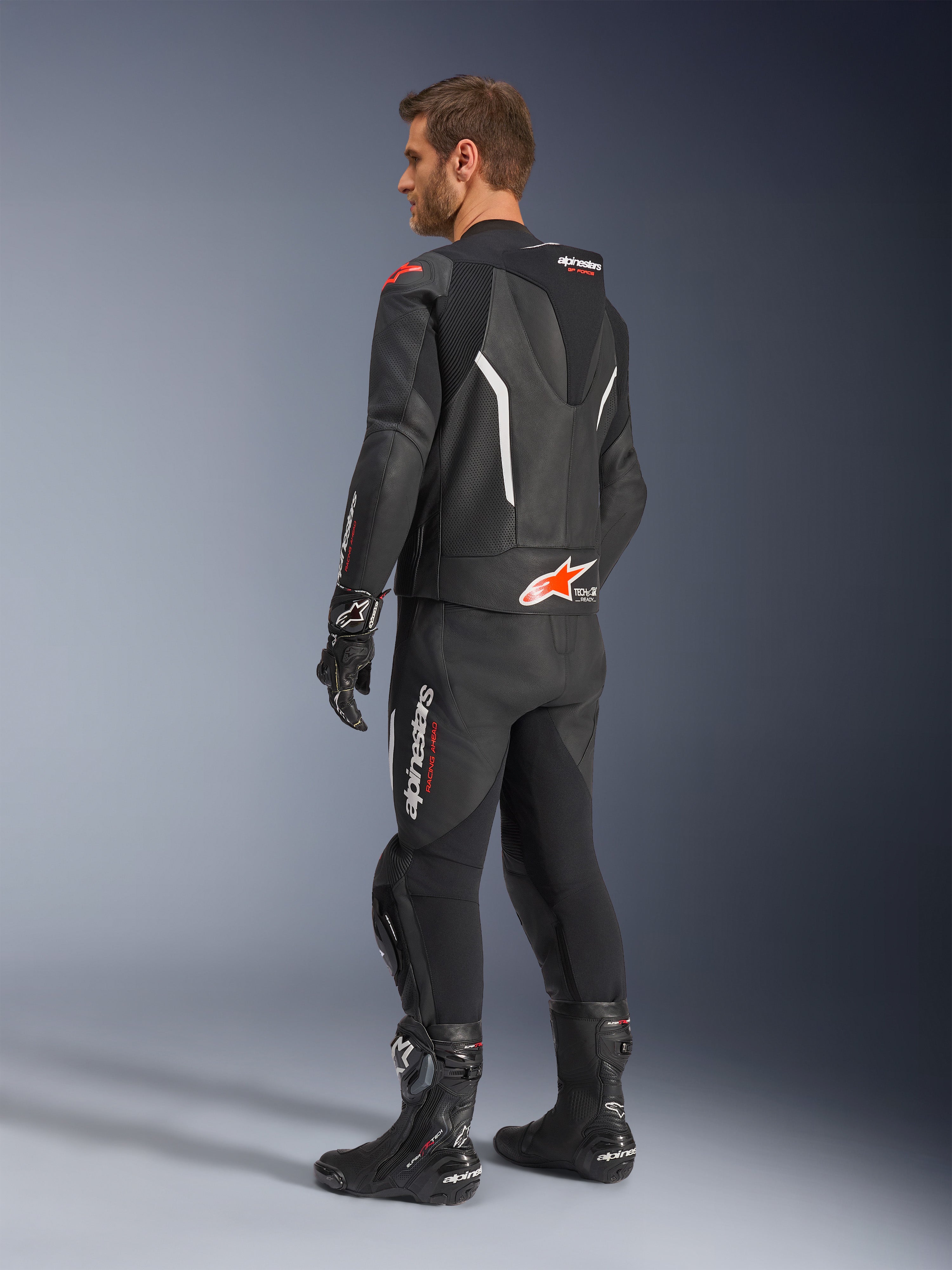 Alpinestars GP Force V2 2-Piece Leather Suit, Black with White and Red Fluo accents, rear three-quarter view on model, featuring an aerodynamic speed hump, perforated leather panels, and bold Alpinestars branding, worn with black racing boots and gloves.