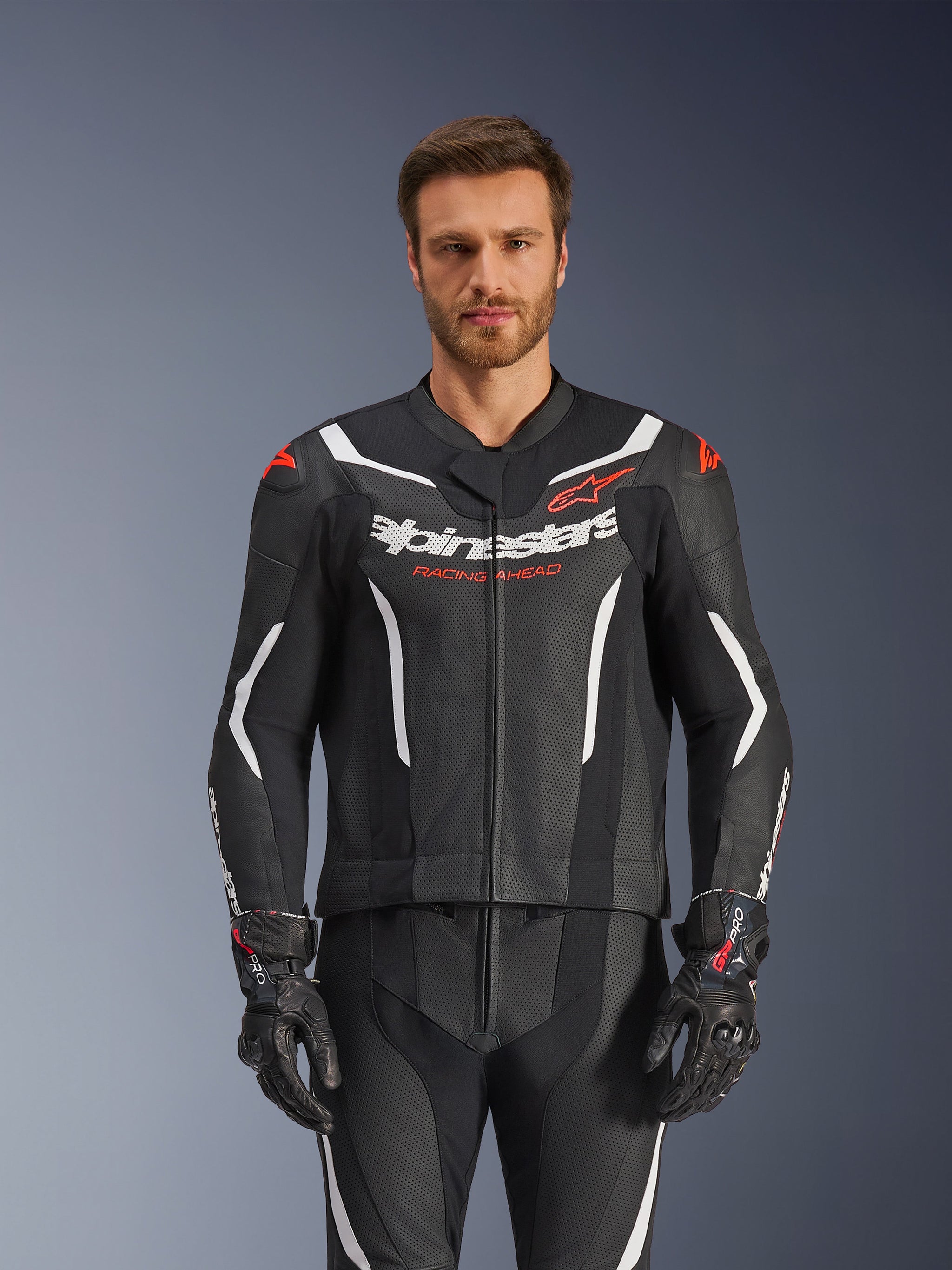 Alpinestars GP Force V2 lederen pak - 2-delig, zwart, wit en fluo rood, vooraanzicht van een mannelijk model dat het geperforeerde lederen pak draagt met bijpassende zwarte handschoenen, met witte hoekige accenten en Alpinestars-branding op de borst