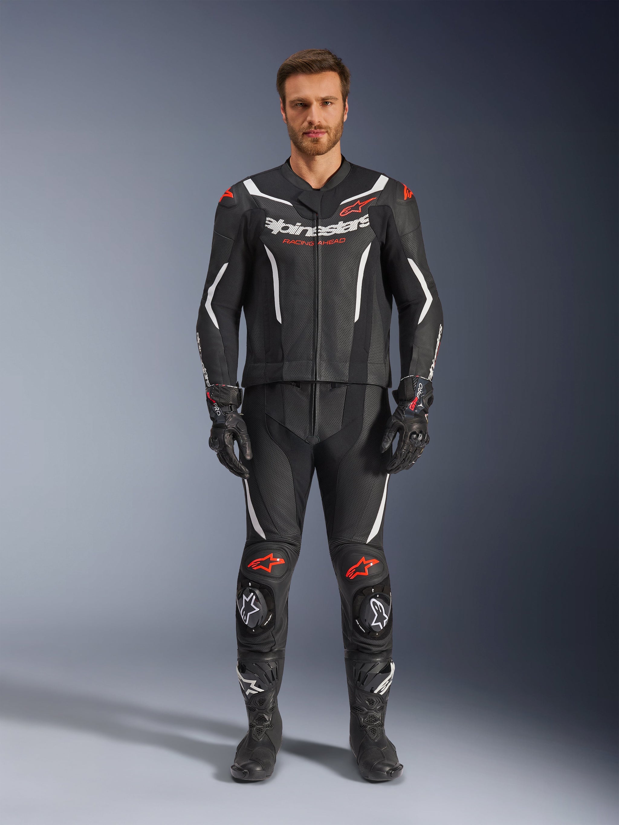 Alpinestars GP Force V2 tweedelig leren pak, motorracepak, zwart, wit en fluo rood, gedragen door een model met bijpassende zwarte handschoenen en laarzen, voorzien van geperforeerde leren panelen, witte en fluo rode accenten en een strak aerodynamisch ontwerp voor circuit- en sportrijden