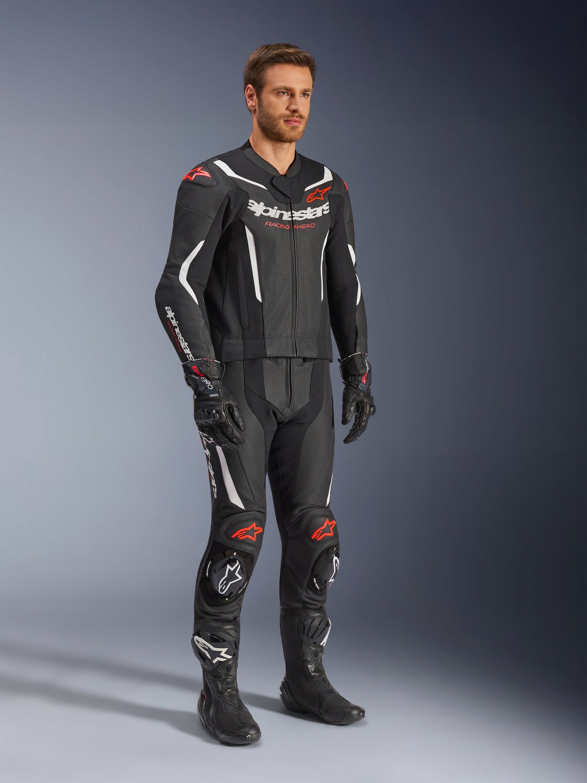 Alpinestars GP Force V2 tweedelig leren pak, zwart, wit en fluo rood, gedragen door een model met zwarte racelaarzen en handschoenen, voorzien van geperforeerde lederen panelen, witte aerodynamische accenten en fluo rode logo's op schouders en knieën, professionele motorrace-uitrusting