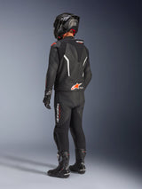 Alpinestars GP Force V2 tweedelig leren pak, zwart motorpak van achteren gezien, gedragen met een bijpassende zwarte helm, handschoenen en laarzen, voorzien van witte en rode accenten, aerodynamische speed hump met Alpinestars-branding, professionele race- en sportuitrusting.