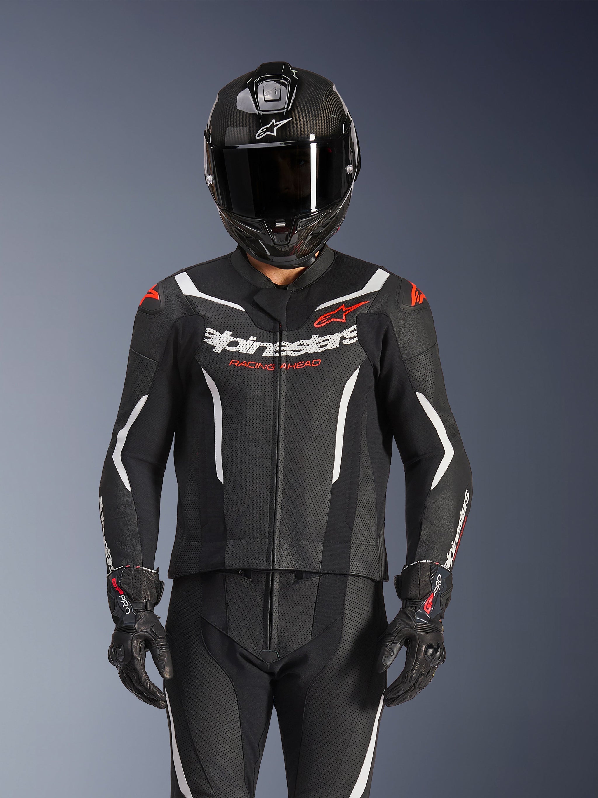 Alpinestars GP Force V2 tweedelig leren pak, zwart wit en fluo rood, gedragen door een rijder met een carbon integraalhelm en zwarte racehandschoenen, voorzien van geperforeerde leren panelen en Alpinestars-branding op de borst.