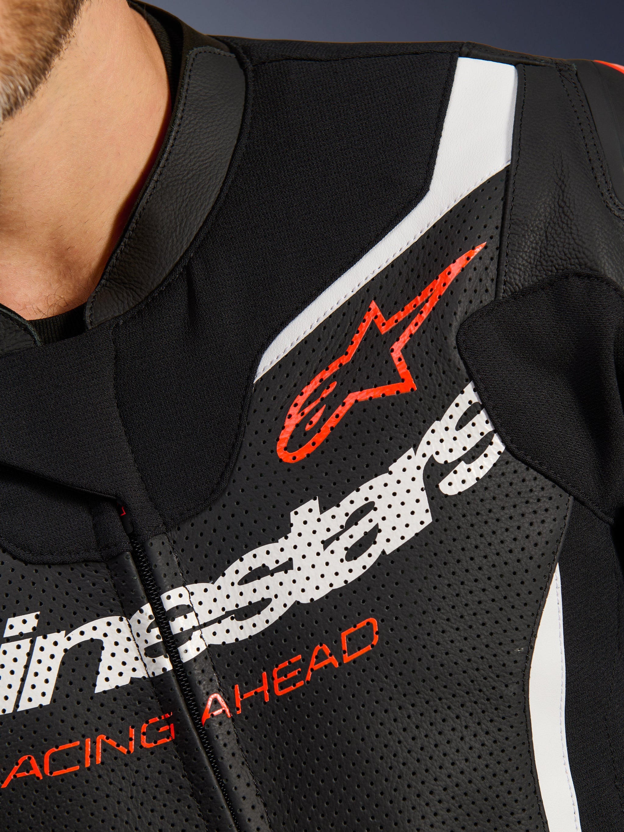 Alpinestars GP Force V2 tweedelig leren pak, close-up van de borst, zwart geperforeerd leer met witte en rode accenten, voorzien van het rode Alpinestars-logo en 'RACING AHEAD'-tekst, detail van hoogwaardige motorracekleding