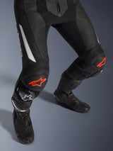 Alpinestars GP Force V2 2-delig leren pak, motorracebroek in zwart met rode en witte accenten, voorzien van geperforeerd leer, accordeon-stretchpanelen boven de knieën met rode sterlogo's, gedragen met zwarte motorlaarzen, hoogwaardige race-uitrusting