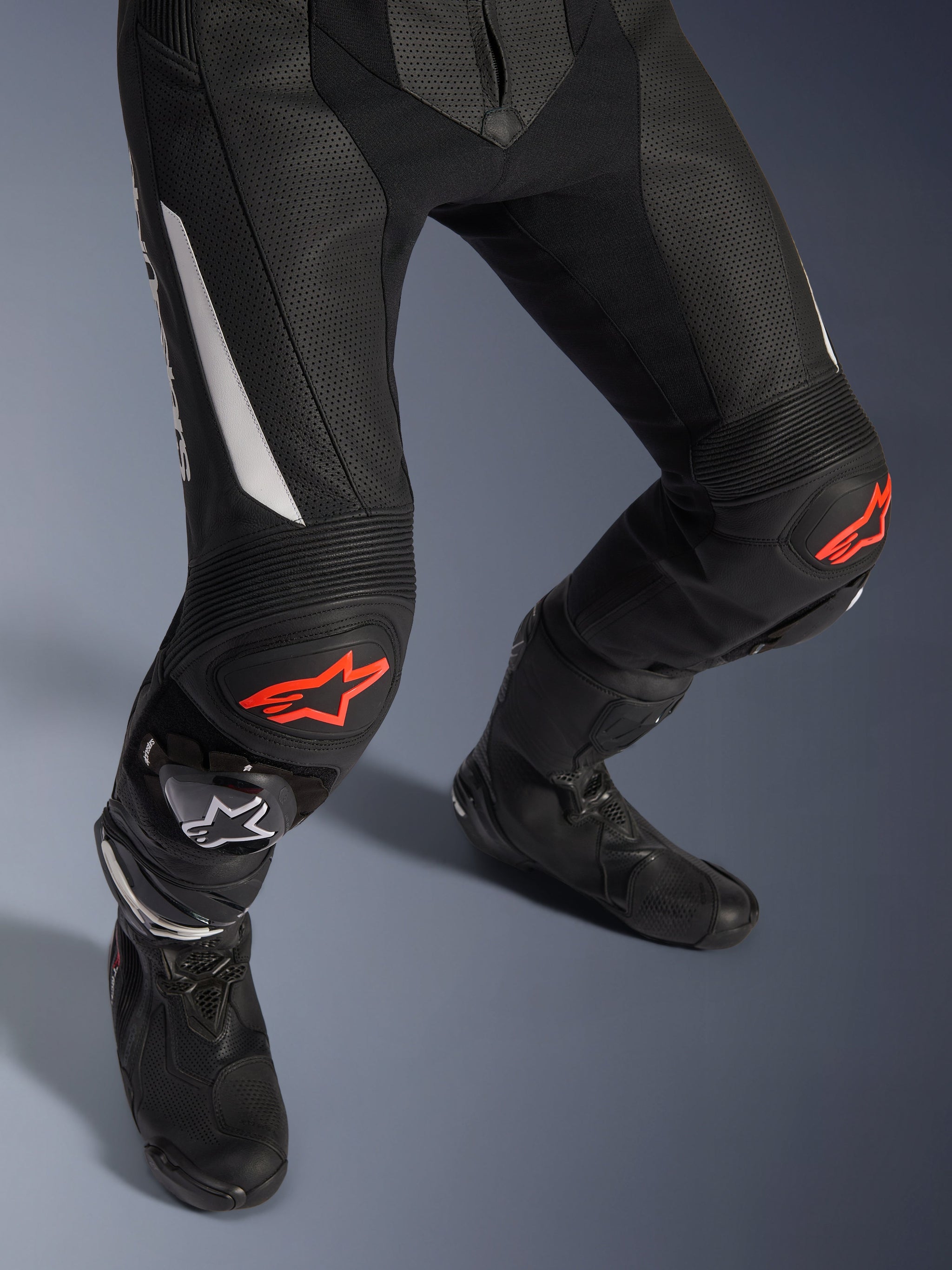 Alpinestars GP Force V2 2-delig leren pak, motorracebroek in zwart met rode en witte accenten, voorzien van geperforeerd leer, accordeon-stretchpanelen boven de knieën met rode sterlogo's, gedragen met zwarte motorlaarzen, hoogwaardige race-uitrusting