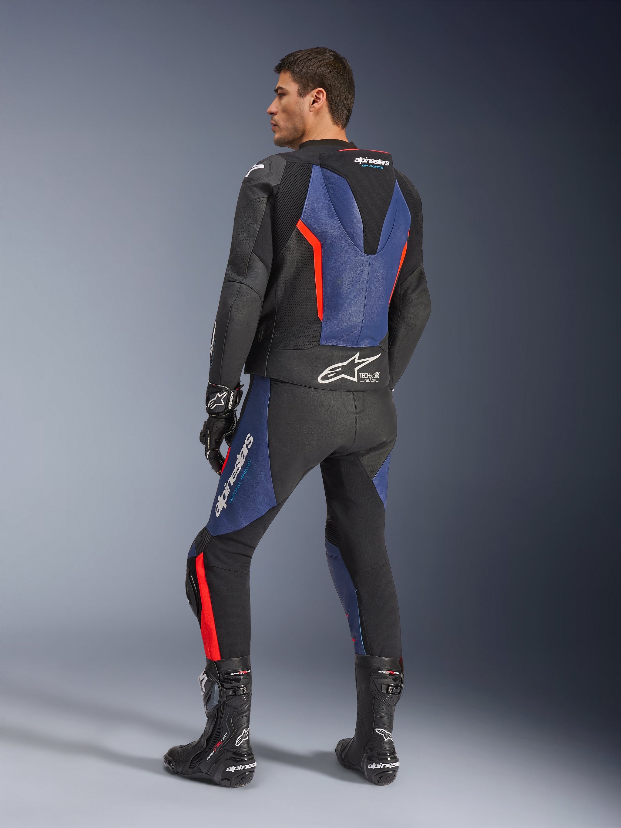 Alpinestars GP Force V2 leren pak (2-delig), zwart, blauw en fluo rood, achteraanzicht op een model, met aerodynamische hump, geperforeerd leer en Tech-Air Ready-branding, gedragen met zwarte Alpinestars racelaarzen en handschoenen.