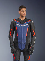 Mannelijk model in een Alpinestars GP Force V2 tweedelig leren pak, zwart blauw rood fluo, met een opvallend blauw borstpaneel en rode accenten, gedragen met GP Pro-handschoenen, professionele leren motorrace-uitrusting met geperforeerde textuur.