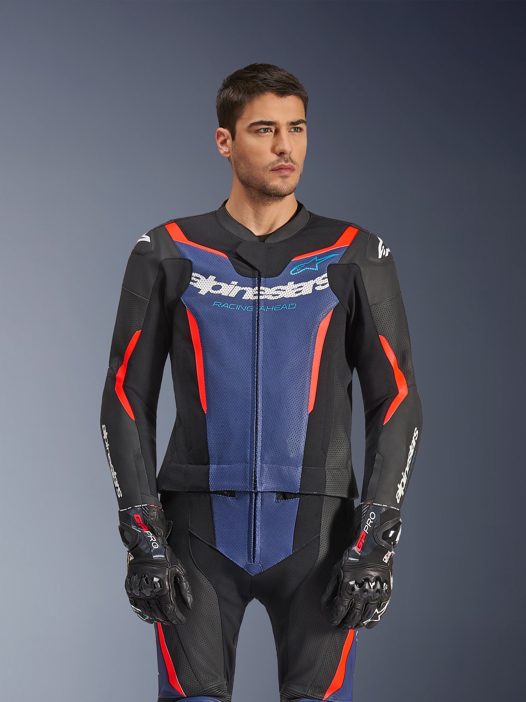 Mannelijk model in een Alpinestars GP Force V2 tweedelig leren pak, zwart blauw rood fluo, met een opvallend blauw borstpaneel en rode accenten, gedragen met GP Pro-handschoenen, professionele leren motorrace-uitrusting met geperforeerde textuur.