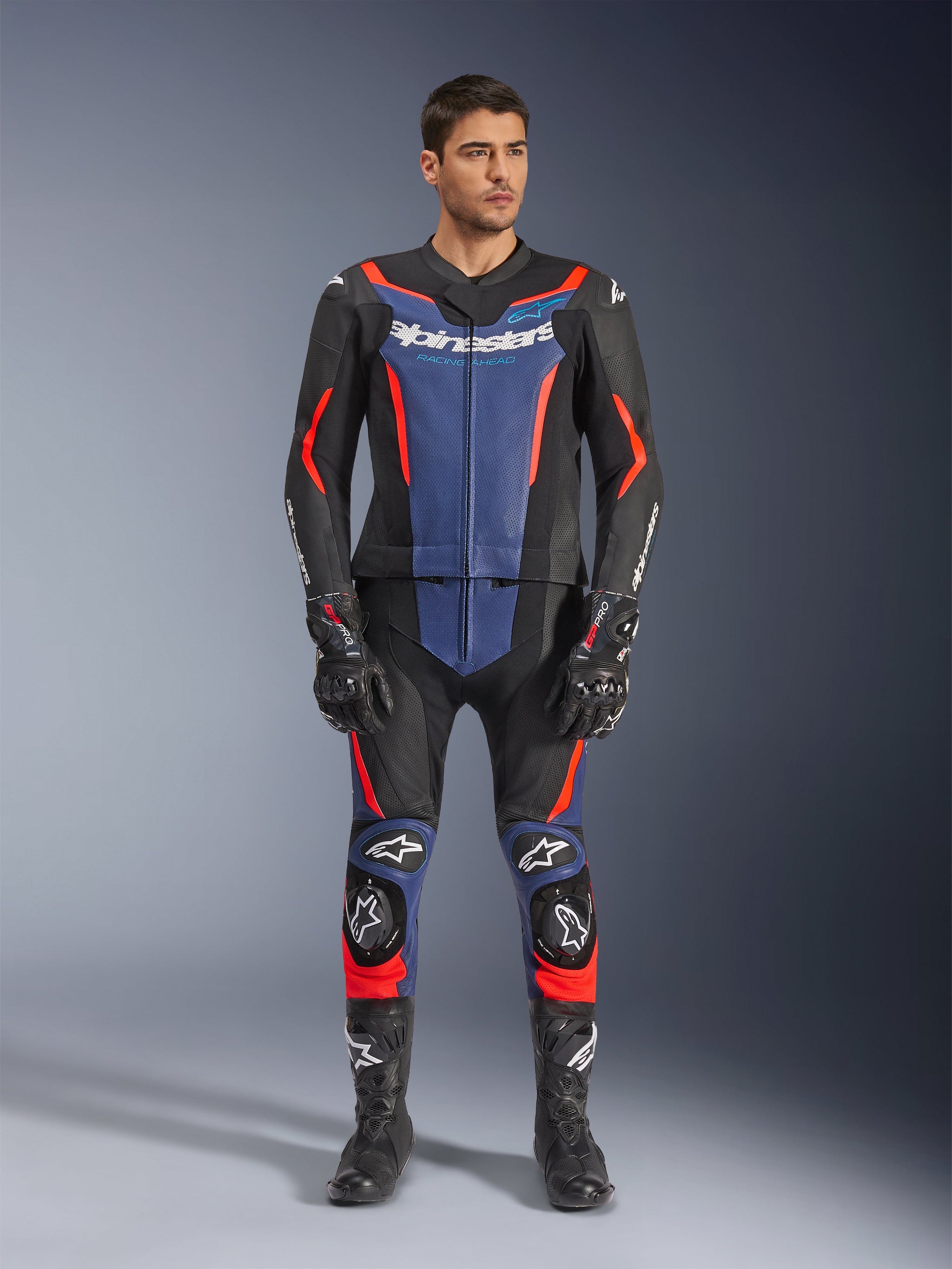 Alpinestars GP Force V2 Leather Suit 2-Piece, tweedelig leren motorpak in zwart, blauw en fluo rood, voorzien van blauwe panelen op de borst en onderbenen met felrode fluorescerende accenten, gedragen door een mannelijk model met zwarte racelaarzen en handschoenen, professionele beschermende kleding voor de motorsport