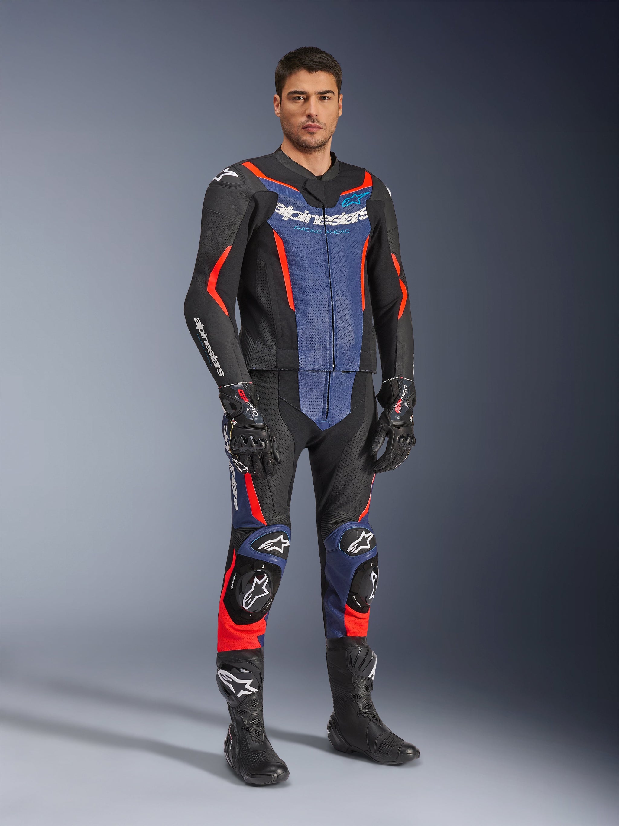 Alpinestars GP Force V2 Leather Suit 2-Piece, 2-delig lederen motorpak, zwart, blauw en fluo rood, gedragen met bijpassende zwarte racehandschoenen en laarzen, voorzien van geperforeerde panelen en beschermende sliders, aerodynamisch ontwerp voor sportief rijden