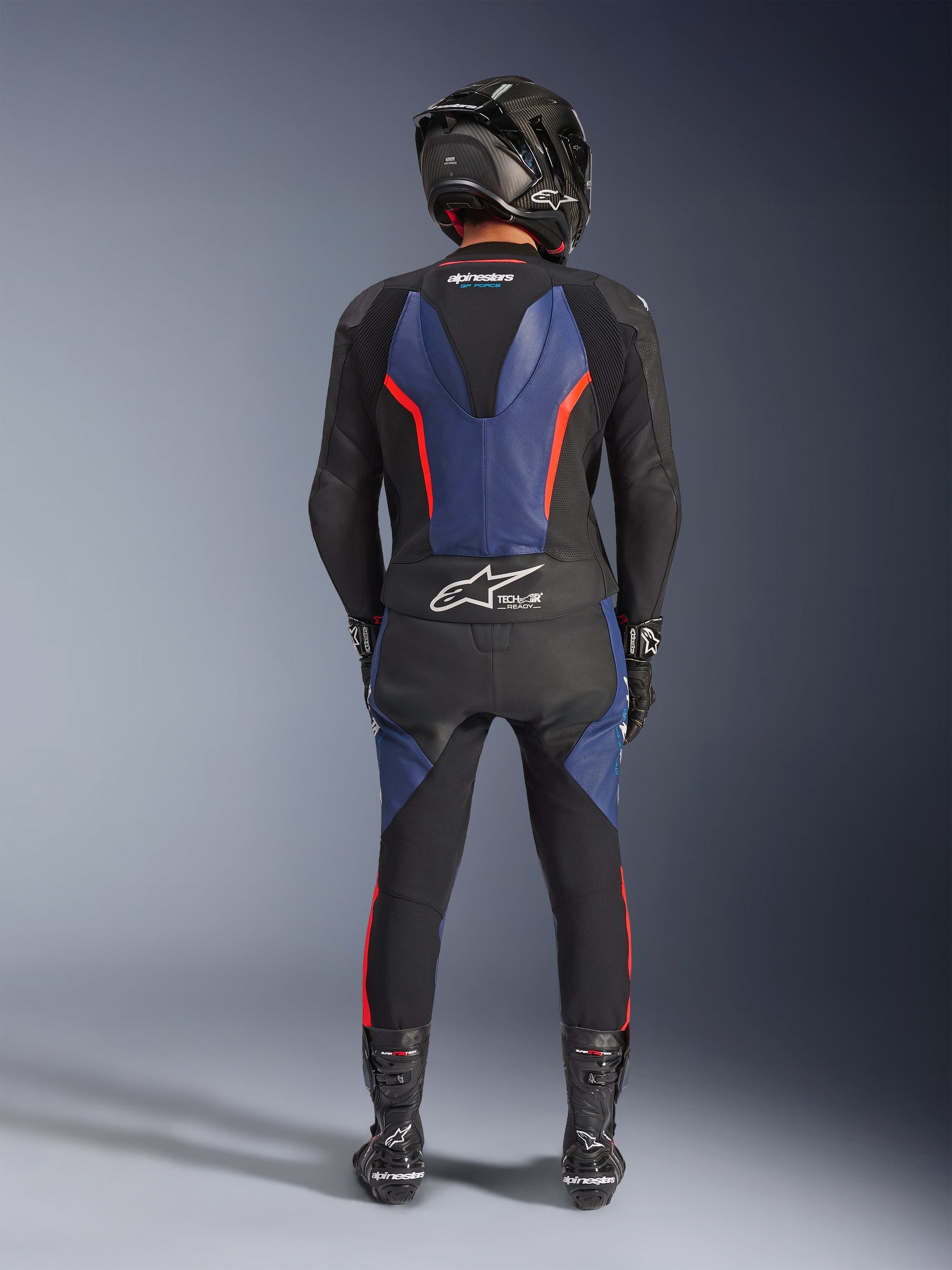 Alpinestars GP Force V2 Leren Pak 2-delig, zwart blauw rood fluo, volledig achteraanzicht van een rijder in complete race-uitrusting, met blauwe lederen panelen op de rug en benen met fluorescerende rode accenten, Tech-Air® Ready-logo op de onderrug, gecombineerd met een carbon helm, zwarte handschoenen en racelaarzen.