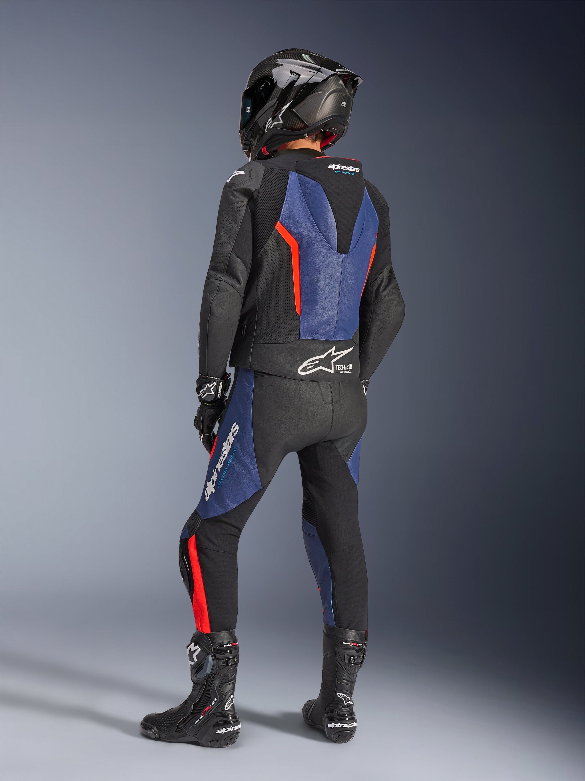 Alpinestars GP Force V2 tweedelig leren motorpak, zwart blauw rood fluo, achteraanzicht van rijder in professionele race-uitrusting, voorzien van een geïntegreerde speedhump en Tech-Air Ready branding, blauwe en rood fluo accenten op zwart leer, gedragen met een carbon fiber helm en zwarte laarzen, hoogwaardige motorkleding