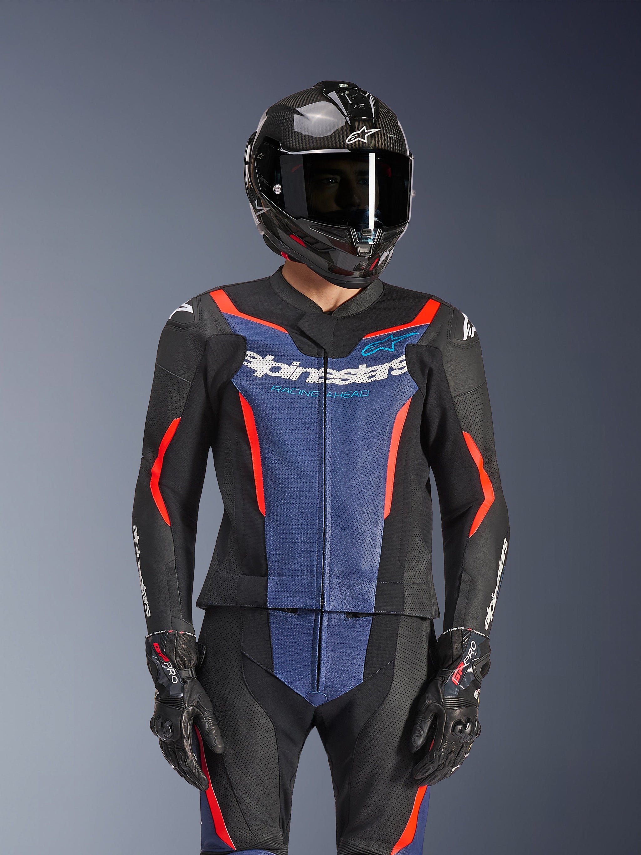 Alpinestars GP Force V2 2-delig leren pak, zwart, blauw en fluo rood, gedragen met een carbon helm en racehandschoenen, voorzien van geperforeerde lederen panelen en wit Alpinestars-logo op de borst, professionele race- en sportuitrusting