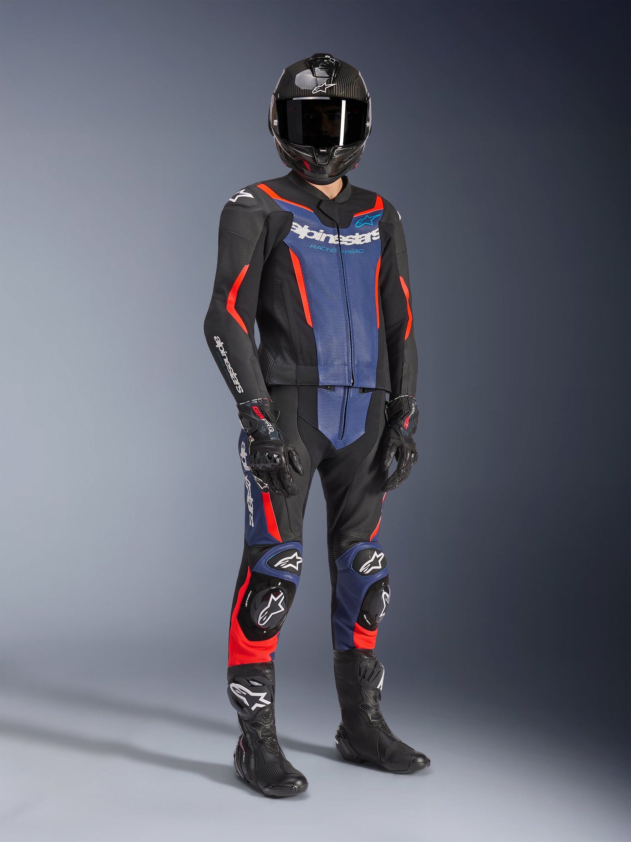 Alpinestars GP Force V2 lederen pak - 2-delig, zwart met blauw en fluo rood, gedragen met een carbon helm, zwarte racehandschoenen en laarzen, voorzien van geperforeerde lederen panelen en wit Alpinestars-logo, professionele sportieve rijuitrusting