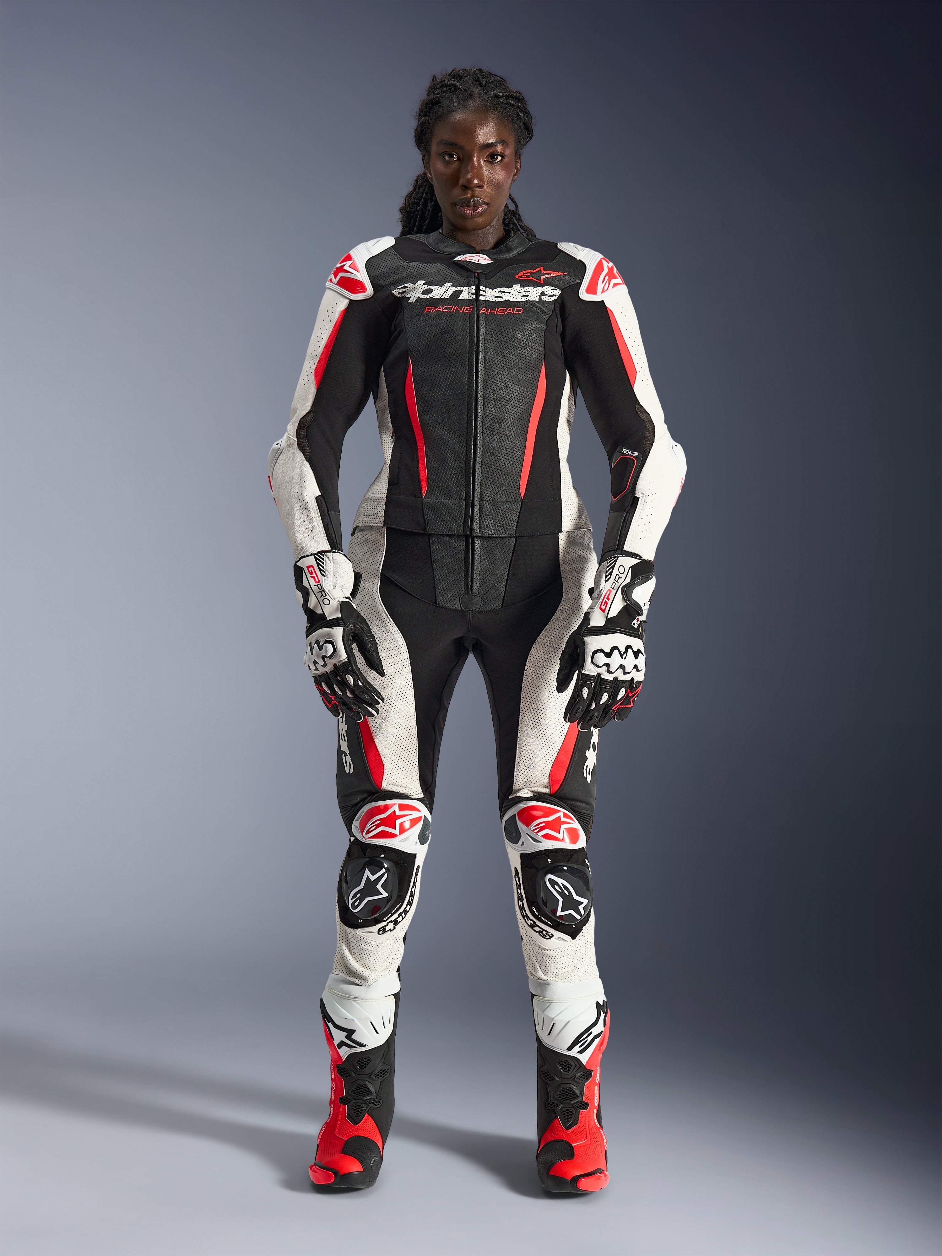 Vrouwelijk model in Alpinestars Stella GP-R7 2-delig leren pak in zwart, wit en Red Fluo, compleet met bijpassende handschoenen en racelaarzen, voorzien van beschermende protectoren en geperforeerde leren panelen, professionele motorrace-uitrusting