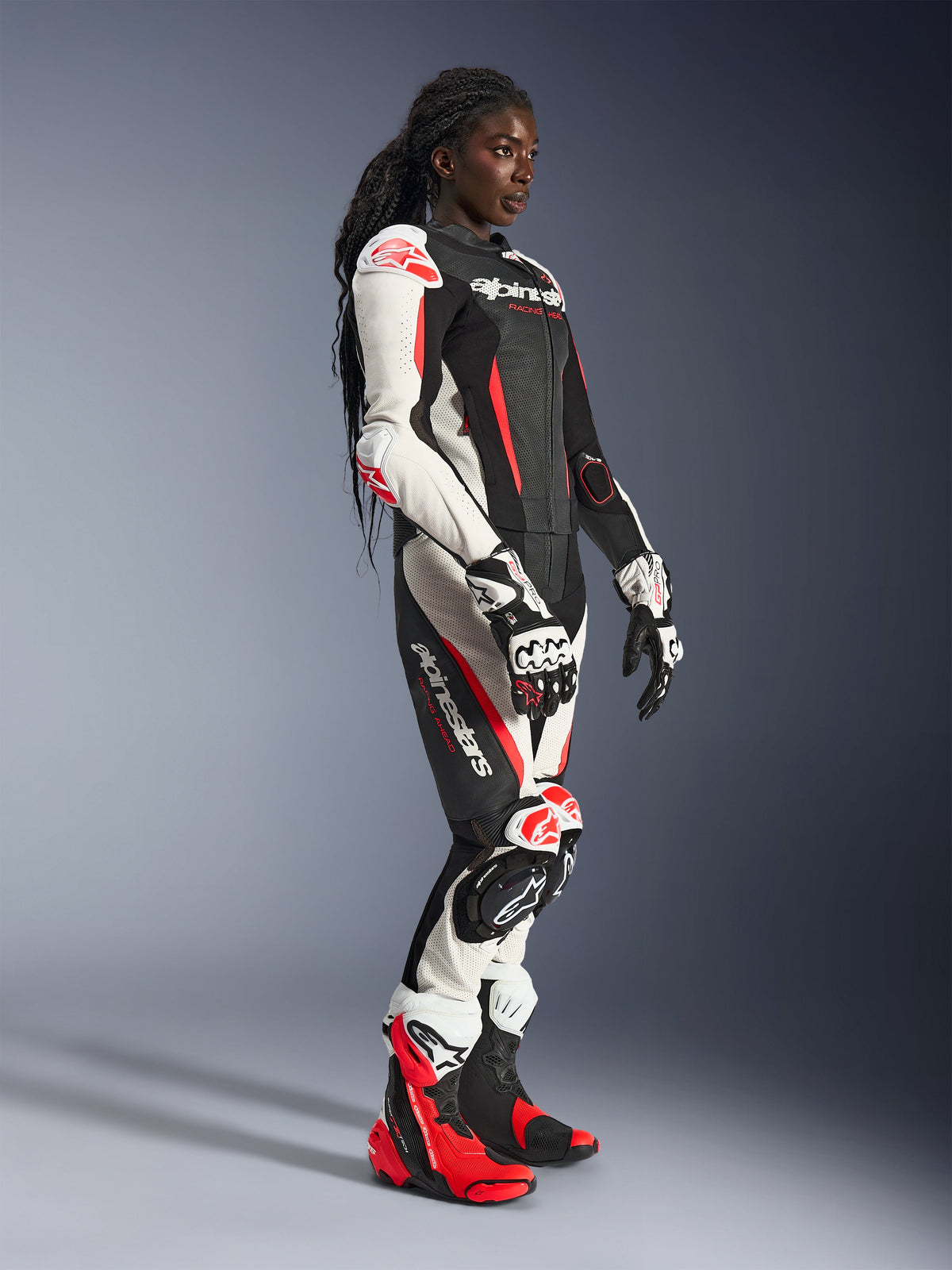 Vrouwelijke rijder in het Alpinestars Stella GP-R7 2-delig leren pak, zwart, wit en fluo rood, gecombineerd met bijpassende racehandschoenen en laarzen, voorzien van geperforeerde leren panelen en protectie op schouders, ellebogen en knieën, staand in een studio-omgeving.