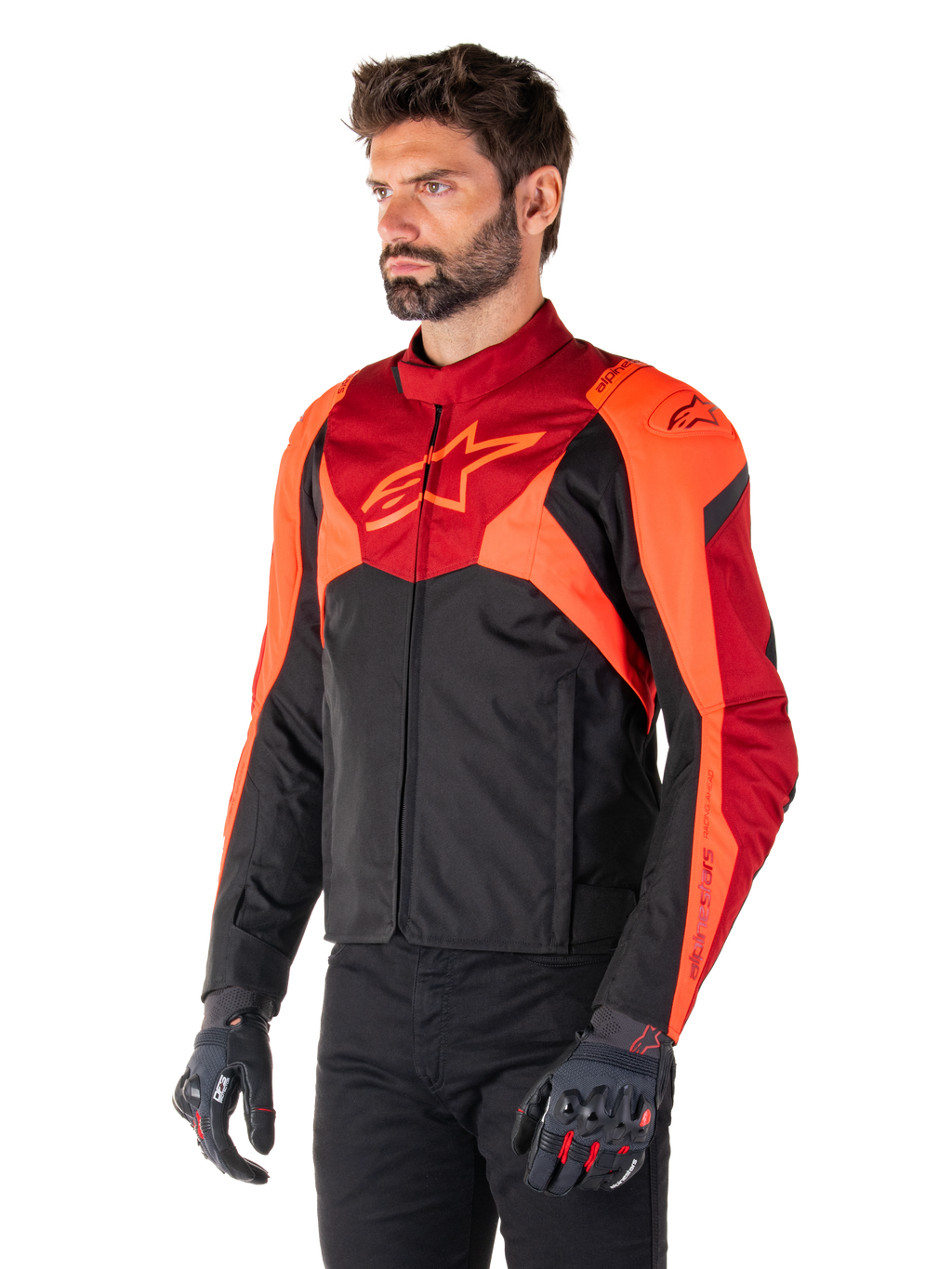 Alpinestars T-Jaws V4 waterdichte jas, zwart, fluo rood en donkerrood, gedragen door een mannelijk model met zwarte broek en motorhandschoenen, agressief sportief silhouet met Alpinestars-logo op de borst, driekwart aanzicht