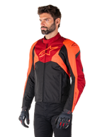 Alpinestars T-Jaws V4 waterdichte jas, zwart, fluo rood en donkerrood, gedragen door een mannelijk model met zwarte broek en motorhandschoenen, agressief sportief silhouet met Alpinestars-logo op de borst, driekwart aanzicht