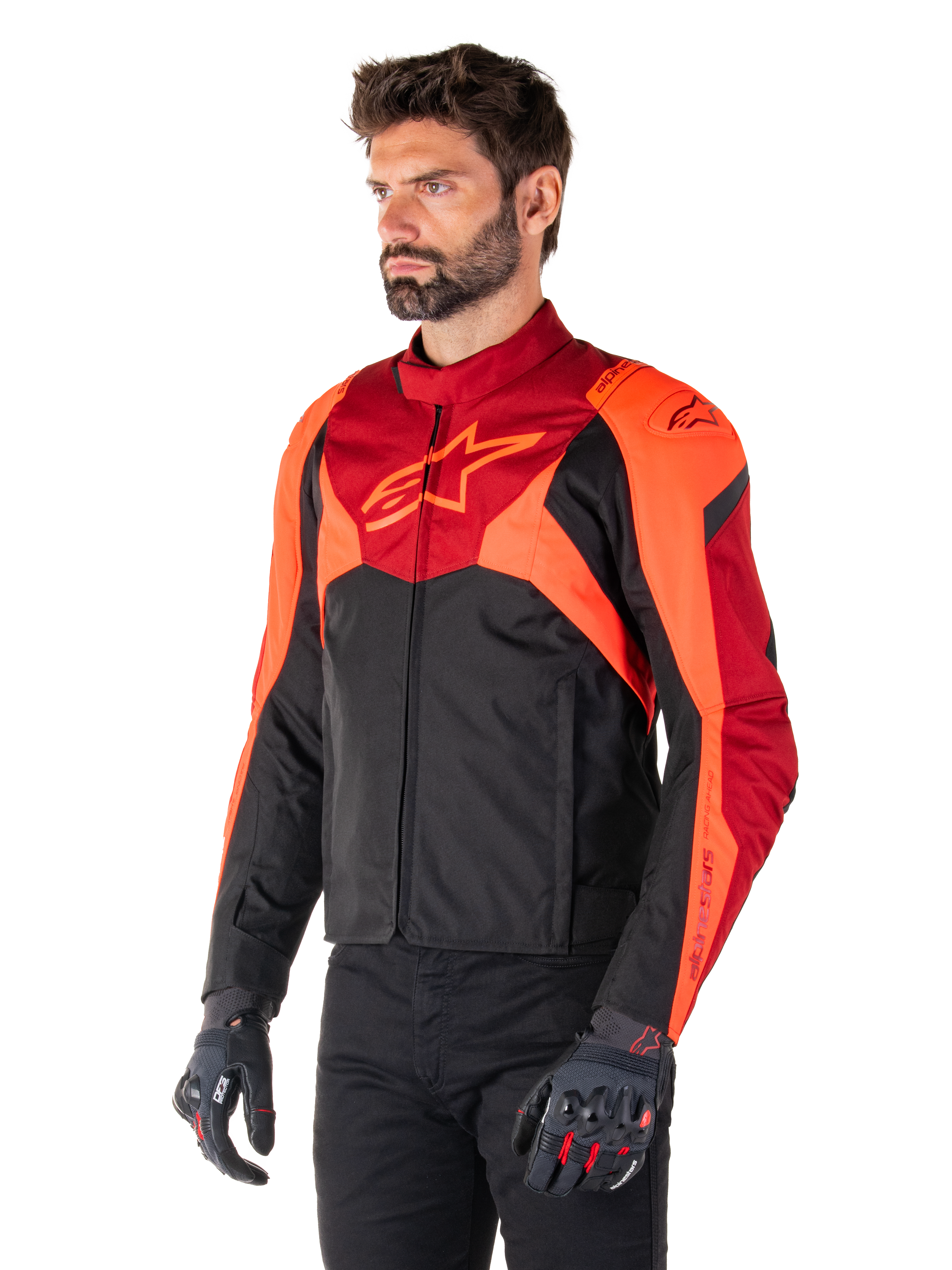 Alpinestars T-Jaws V4 waterdichte jas, zwart, fluo rood en donkerrood, gedragen door een mannelijk model met zwarte broek en motorhandschoenen, agressief sportief silhouet met Alpinestars-logo op de borst, driekwart aanzicht