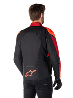 Achteraanzicht van een man die de Alpinestars T-Jaws V4 Waterproof Jas draagt in Black Red Fluo Dark Red, met opvallende rode accenten op de schouders en mouwen, een fluo rood Alpinestars-logo op de onderrug, gecombineerd met een zwarte broek en handschoenen.