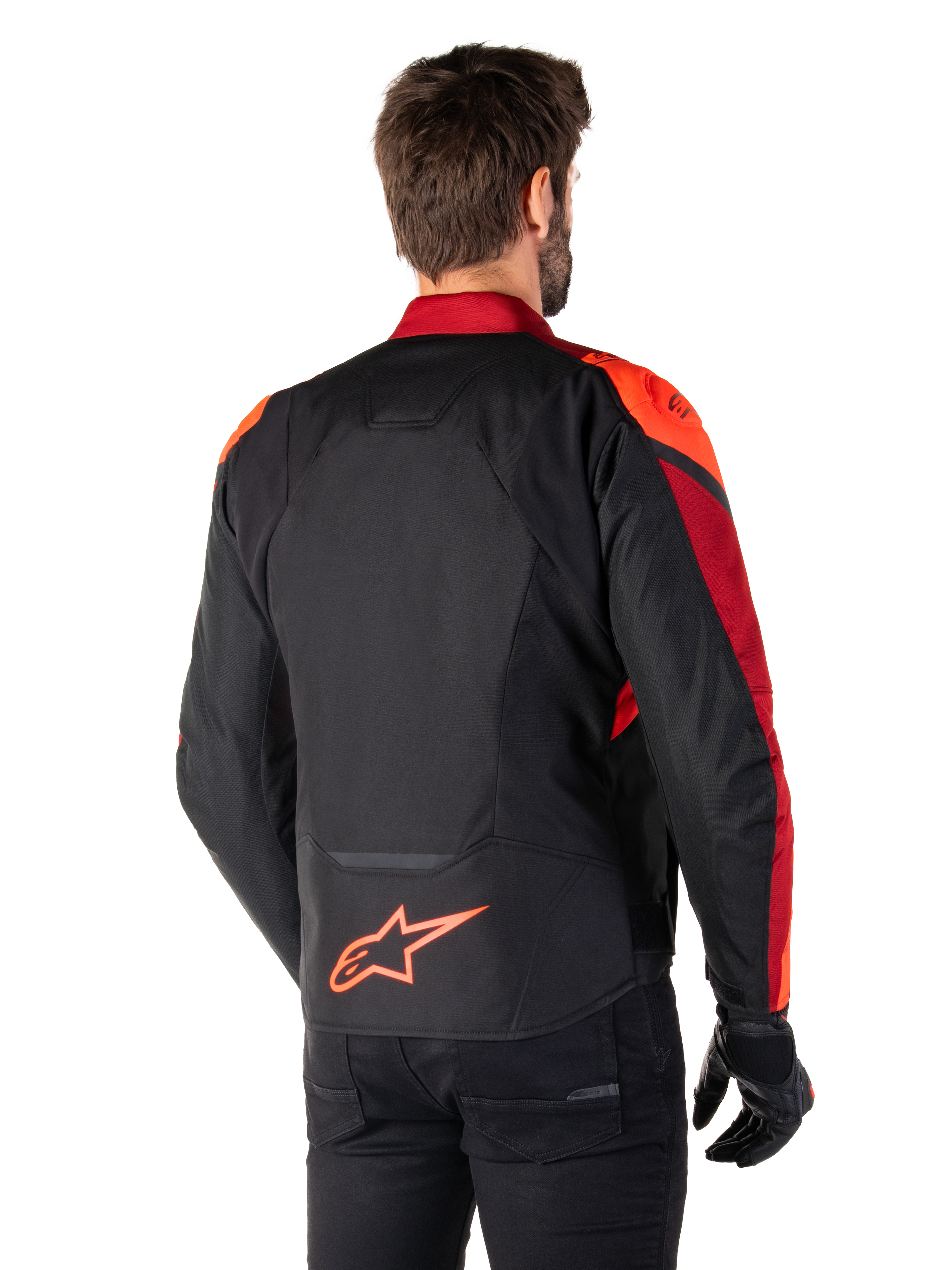 Achteraanzicht van een man die de Alpinestars T-Jaws V4 Waterproof Jas draagt in Black Red Fluo Dark Red, met opvallende rode accenten op de schouders en mouwen, een fluo rood Alpinestars-logo op de onderrug, gecombineerd met een zwarte broek en handschoenen.