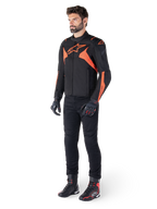 Alpinestars T-Jaws V4 waterdichte jas, zwart en fluo rood, gedragen door een mannelijk model met zwarte broek, handschoenen en laarzen, voorzien van felle oranje accenten en het Alpinestars-logo, sportief ontwerp voor motorrijden