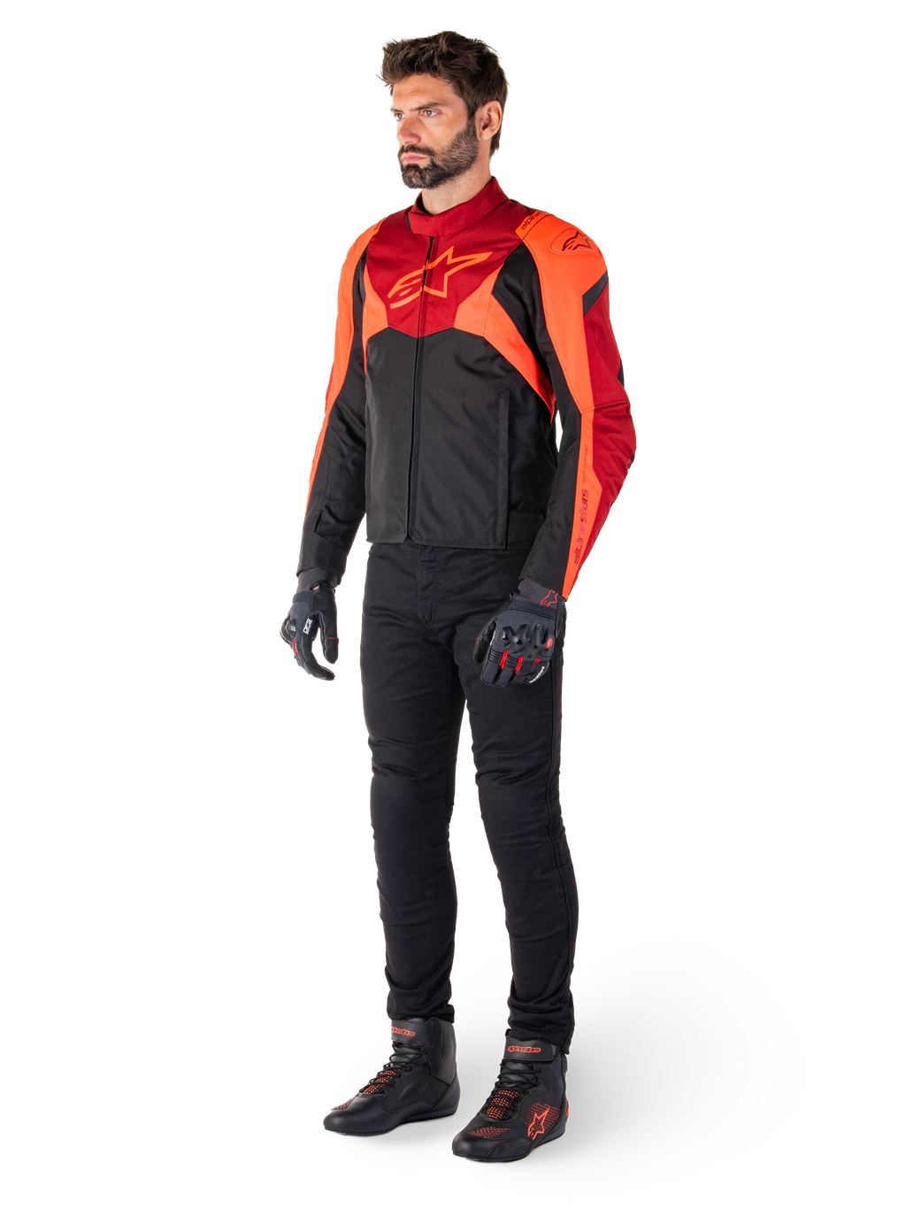 Mannelijke motorrijder in de Alpinestars T-Jaws V4 Waterproof motorjas in zwart, fluo rood en donkerrood, gecombineerd met een zwarte motorbroek, handschoenen en motorlaarzen, voorzien van een sportieve pasvorm, opvallende branding op de borst en een agressief silhouet.