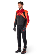 Mannelijke motorrijder in de Alpinestars T-Jaws V4 Waterproof motorjas in zwart, fluo rood en donkerrood, gecombineerd met een zwarte motorbroek, handschoenen en motorlaarzen, voorzien van een sportieve pasvorm, opvallende branding op de borst en een agressief silhouet.
