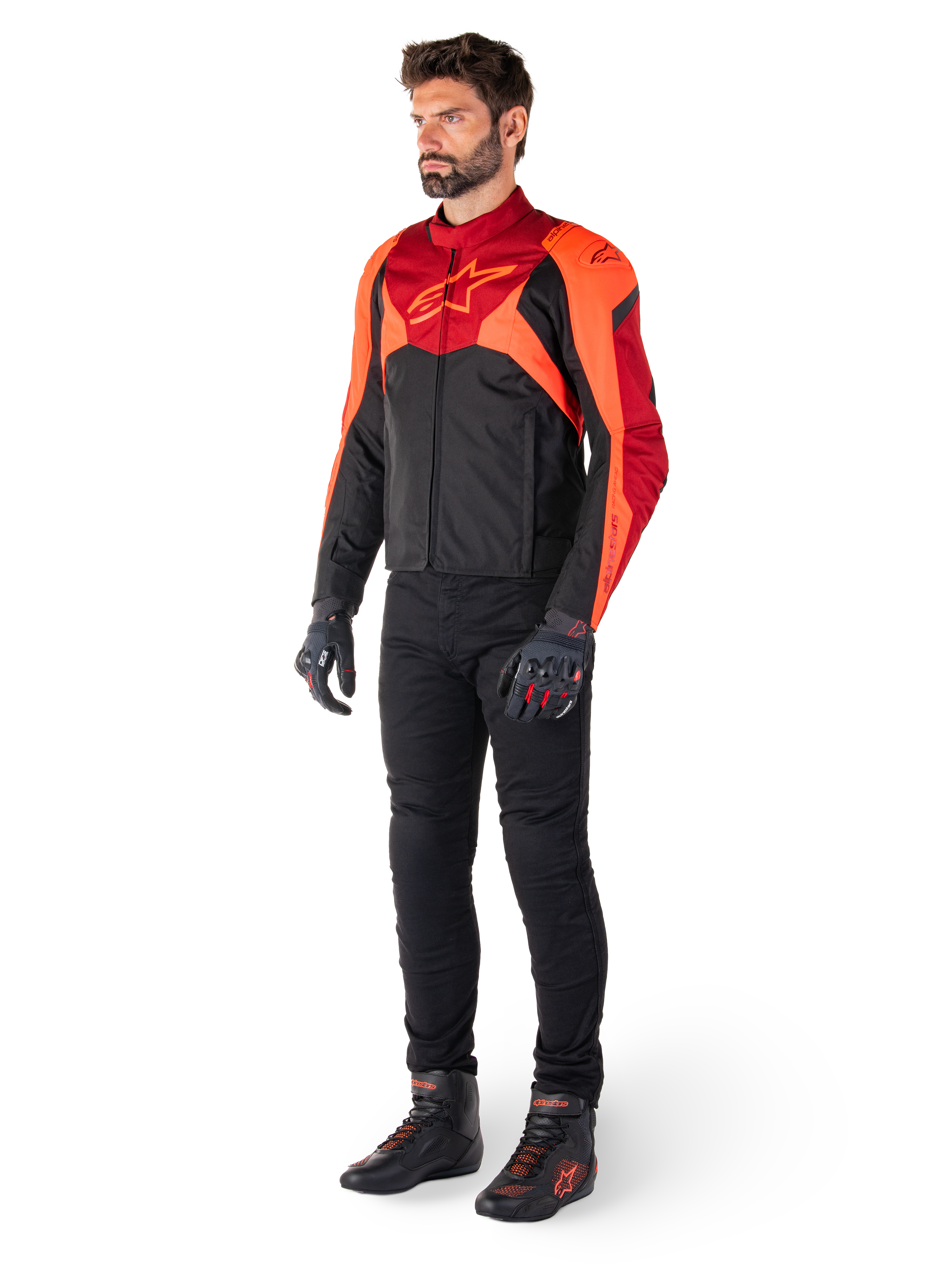 Mannelijke motorrijder in de Alpinestars T-Jaws V4 Waterproof motorjas in zwart, fluo rood en donkerrood, gecombineerd met een zwarte motorbroek, handschoenen en motorlaarzen, voorzien van een sportieve pasvorm, opvallende branding op de borst en een agressief silhouet.