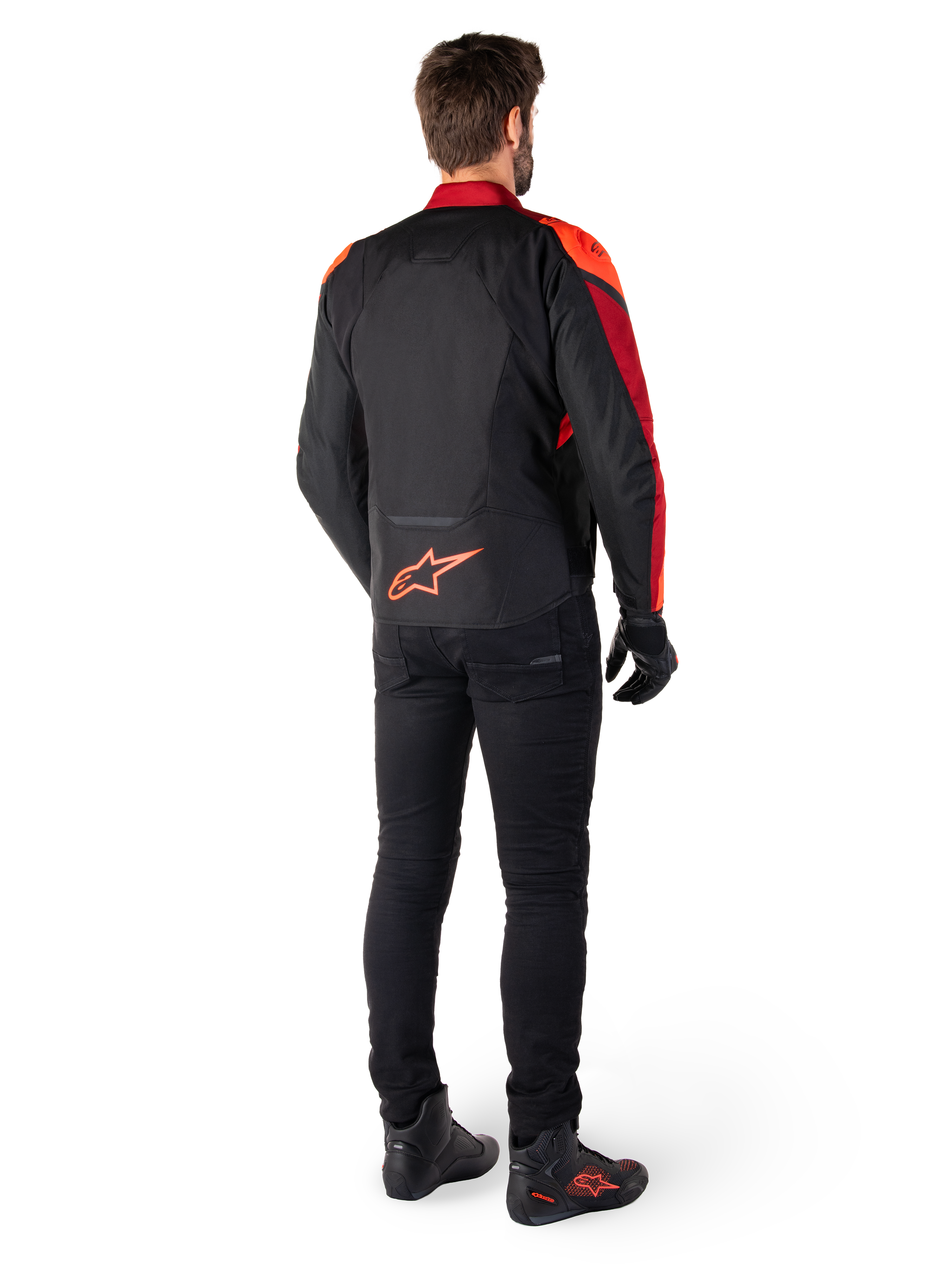 Alpinestars T-Jaws V4 Waterproof Jas in Black Red Fluo Dark Red, achteraanzicht van een motorrijder die de textiele jas draagt met een zwarte broek en motorlaarzen, voorzien van een opvallend rood Alpinestars-logo op de onderrug.