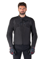Alpinestars T-Jaws V4 Waterproof Jacket, zwarte sportieve motorjas, gedragen door een man met zwarte handschoenen met rode accenten, versterkte schouders en armen, gestroomlijnd design met subtiel logo op de borst, textiele all-weather motorkleding