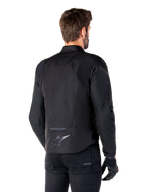 Alpinestars T-Jaws V4 Waterproof Jacket, zwart, achteraanzicht op model, met een reflecterend Alpinestars-logo op de onderrug, verlengde achterkant voor extra dekking, sportief textielontwerp met tonale panelen, gedragen met zwarte motorhandschoenen