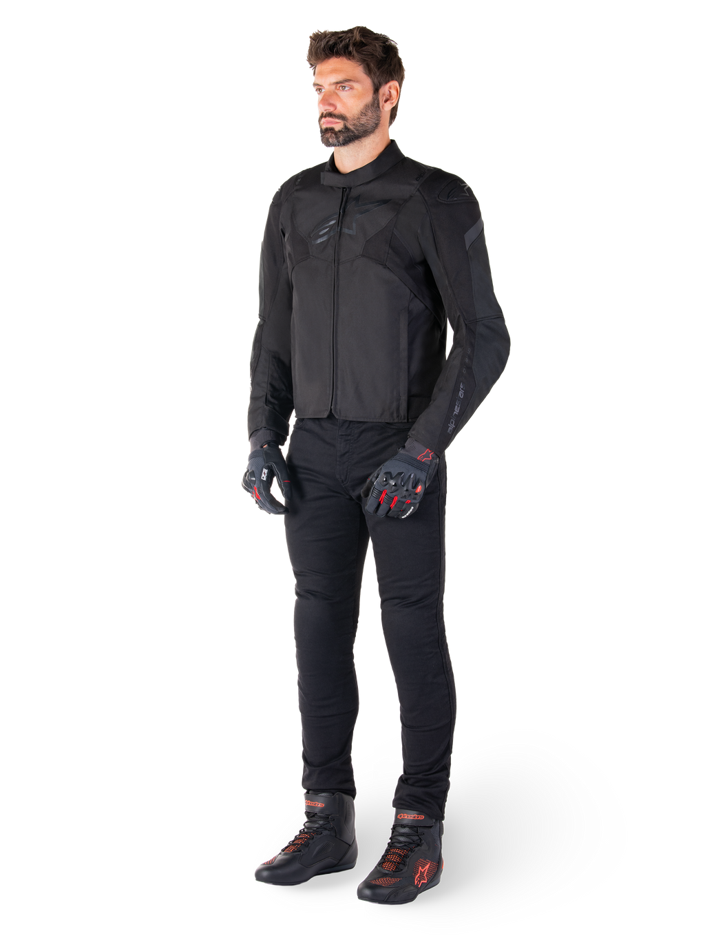 Mannelijk model draagt de Alpinestars T-Jaws V4 Waterdichte Jas in het zwart, gecombineerd met een zwarte motorbroek, zwarte handschoenen met rode accenten en zwarte motorschoenen, met een slanke sportieve pasvorm en subtiele branding, geïsoleerd op een witte achtergrond.
